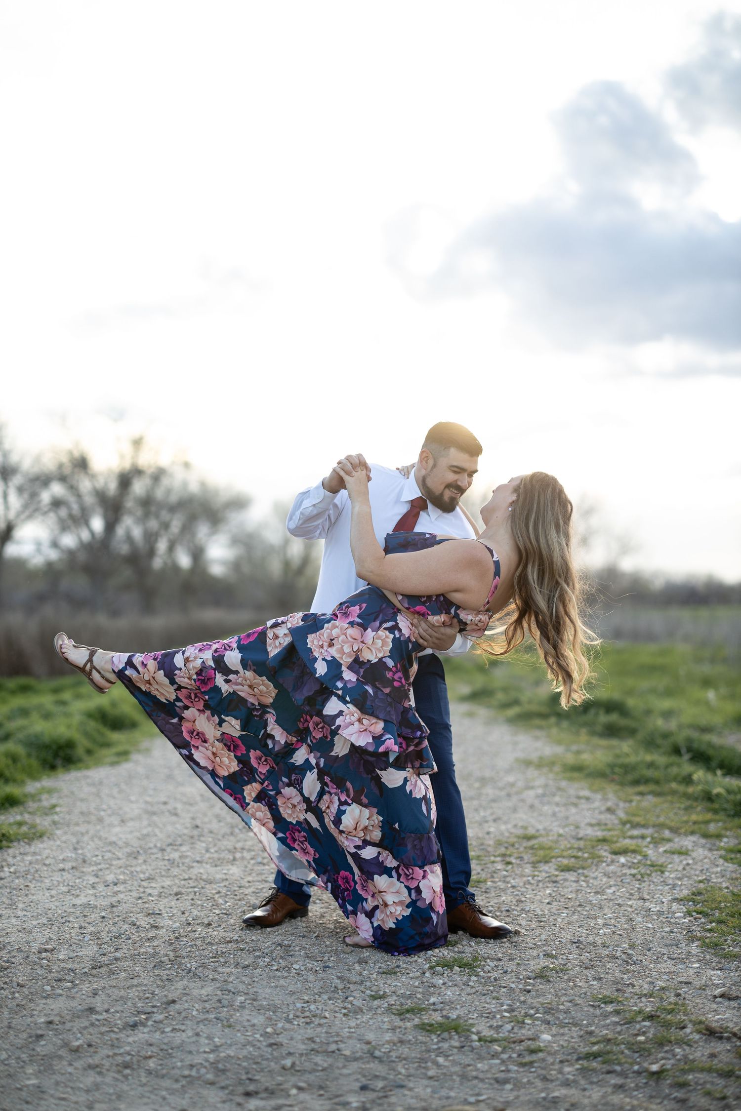 Dynamic dance dip pose on a gravel path surrounded by green fields.