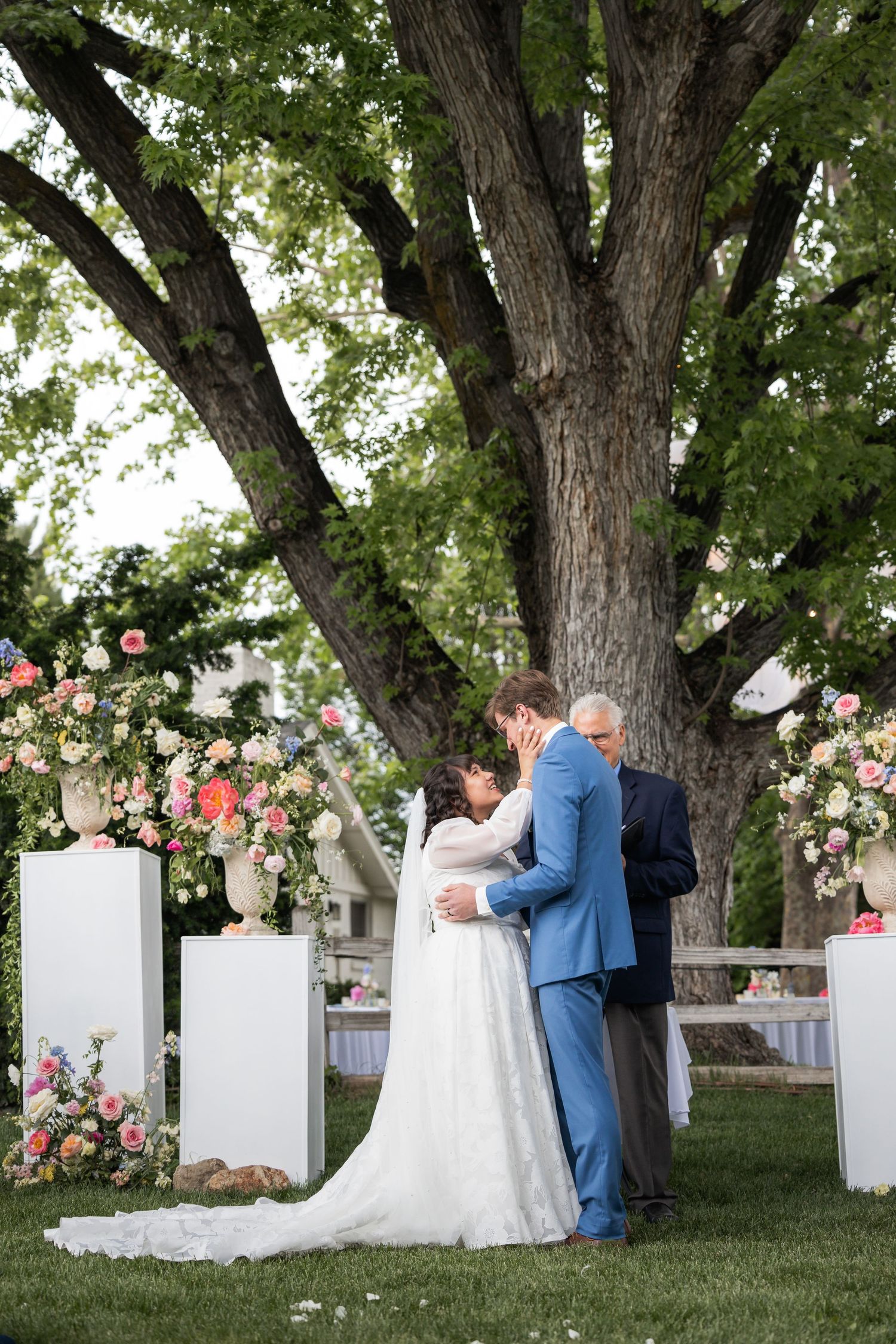 Romantic wedding ceremony under large tree with floral decorations and white pedestals.