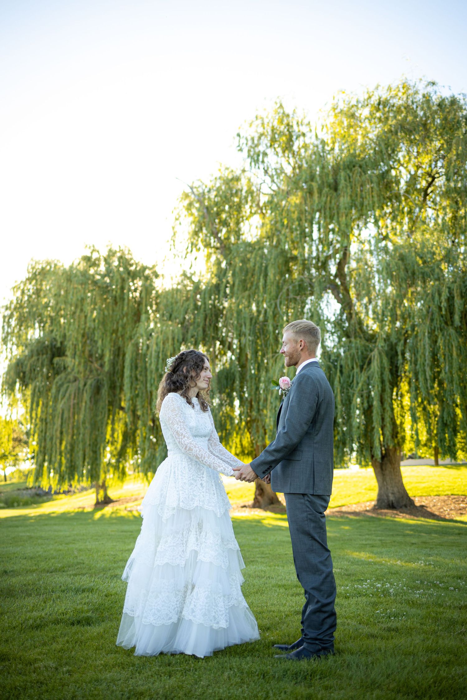 Wedding couple stands together on grass with majestic weeping willow trees in background.