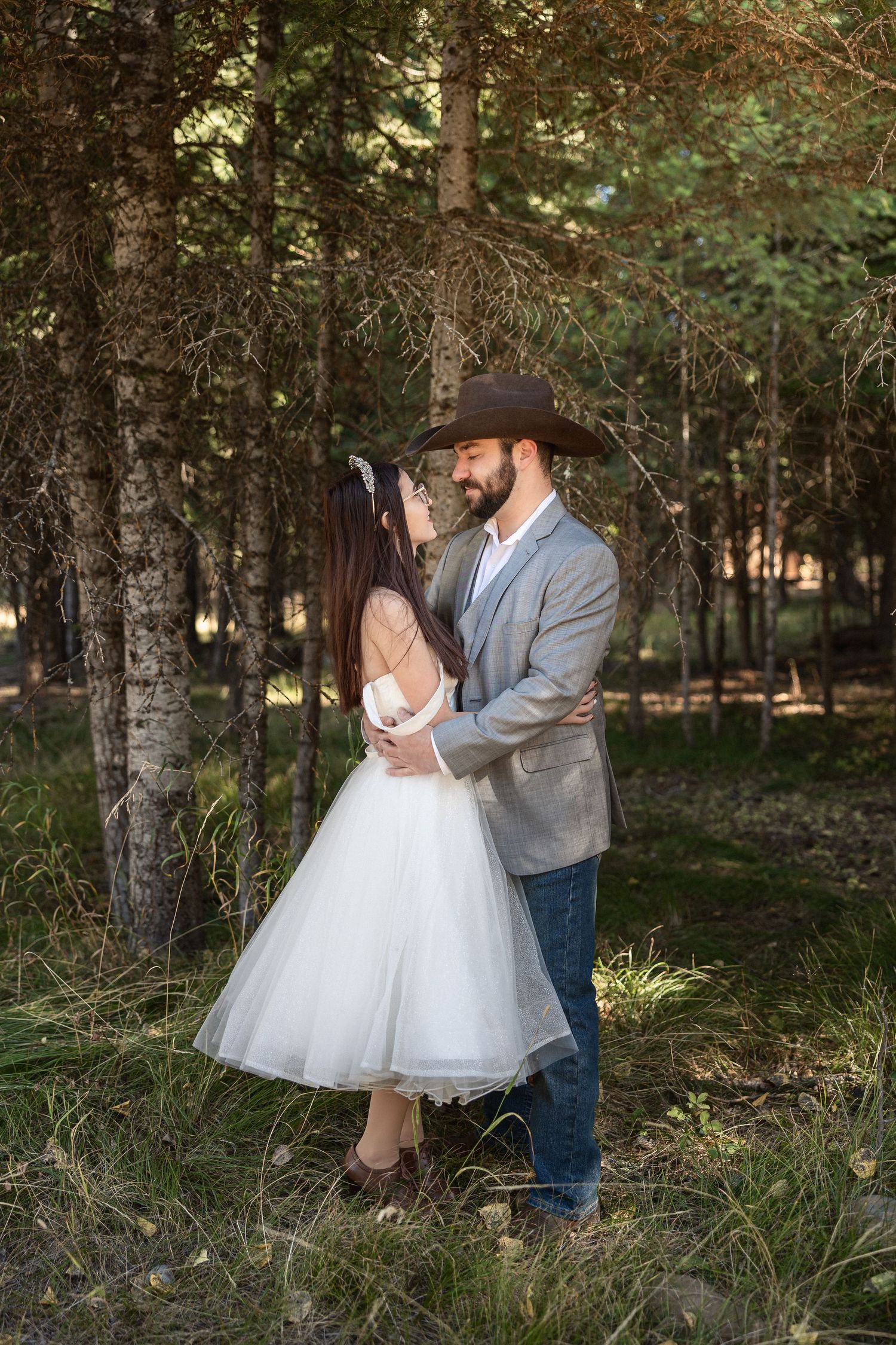 A romantic wedding couple embraces in a forest setting with the bride wearing a tea-length white dress.