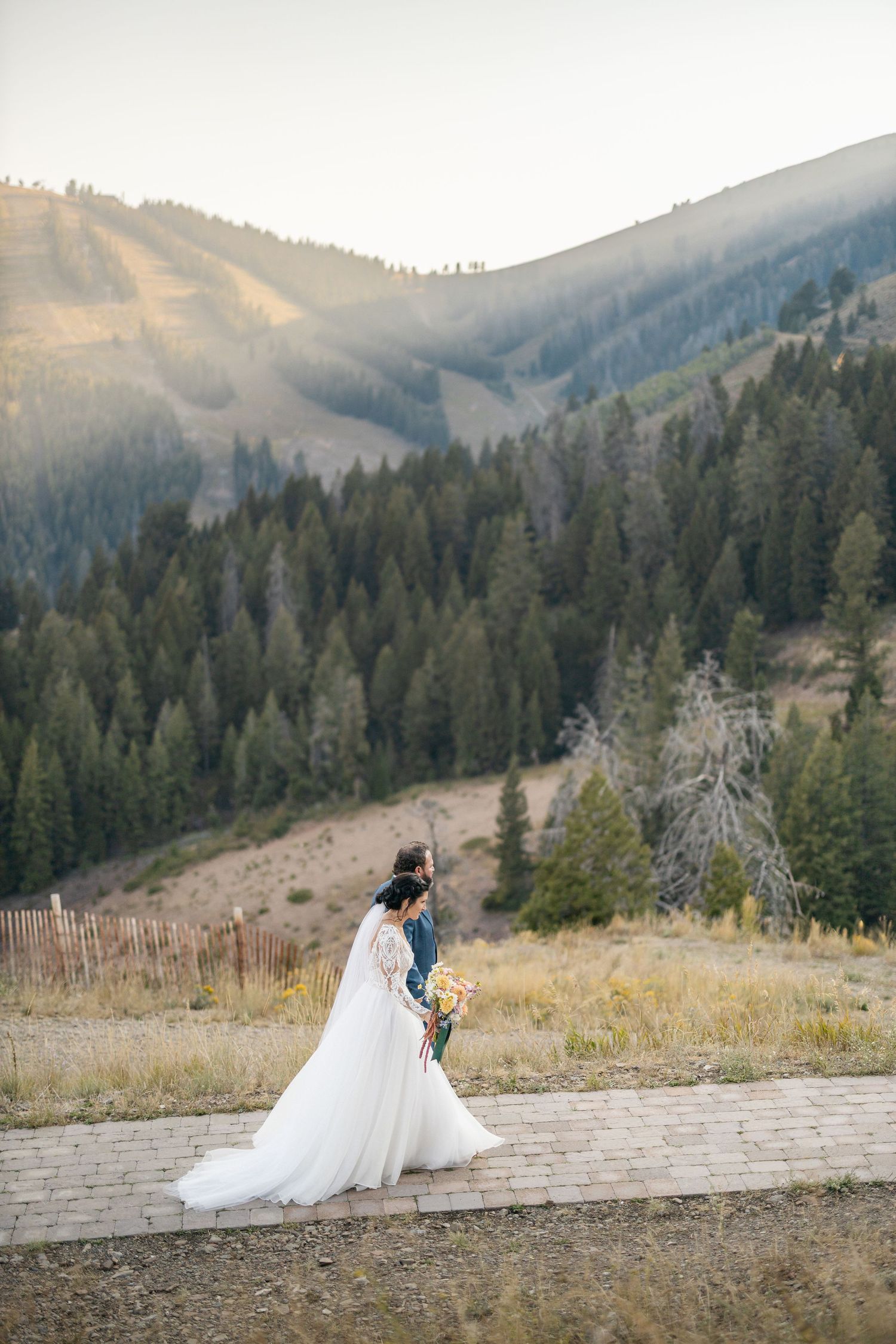 A wedding couple poses on a mountain path with evergreen forests and rolling hills in the background.