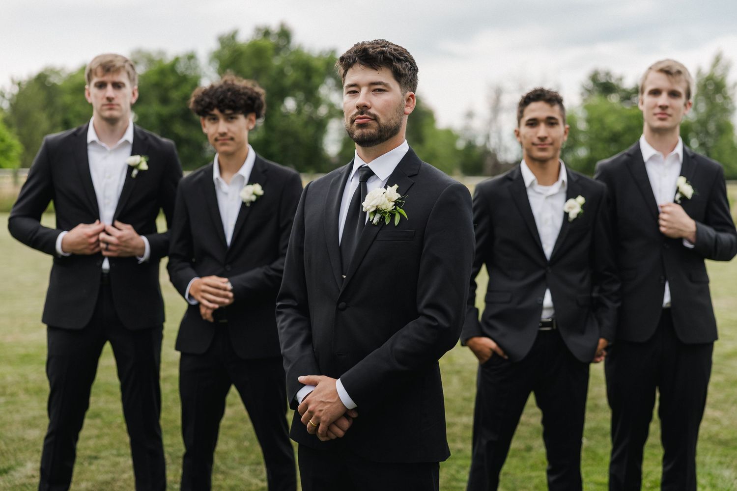 Groom in black suit with white boutonniere stands before four groomsmen outdoors at a wedding ceremony.