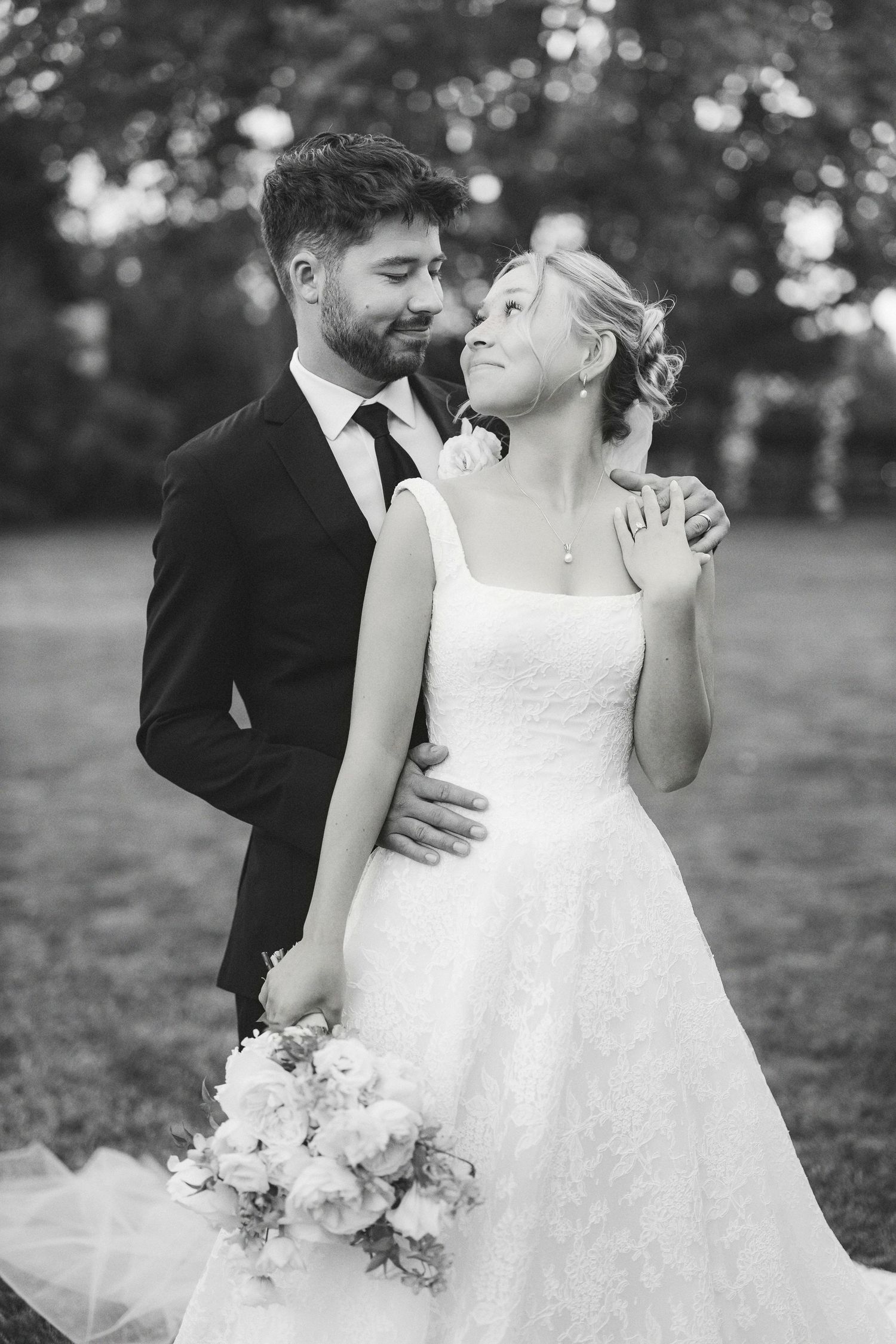 Black and white photo of a smiling bride holding flowers, embraced by groom in suit outdoors.