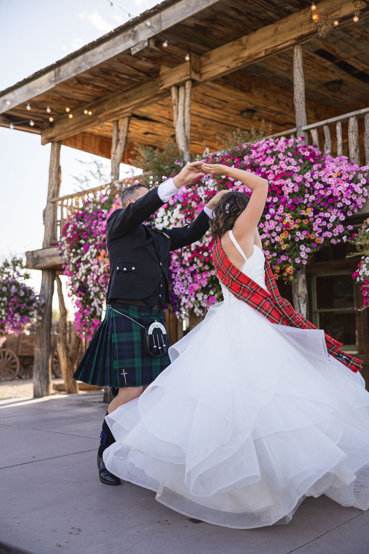 Wedding couple dancing playfully under rustic wooden porch with blooming pink flowers.