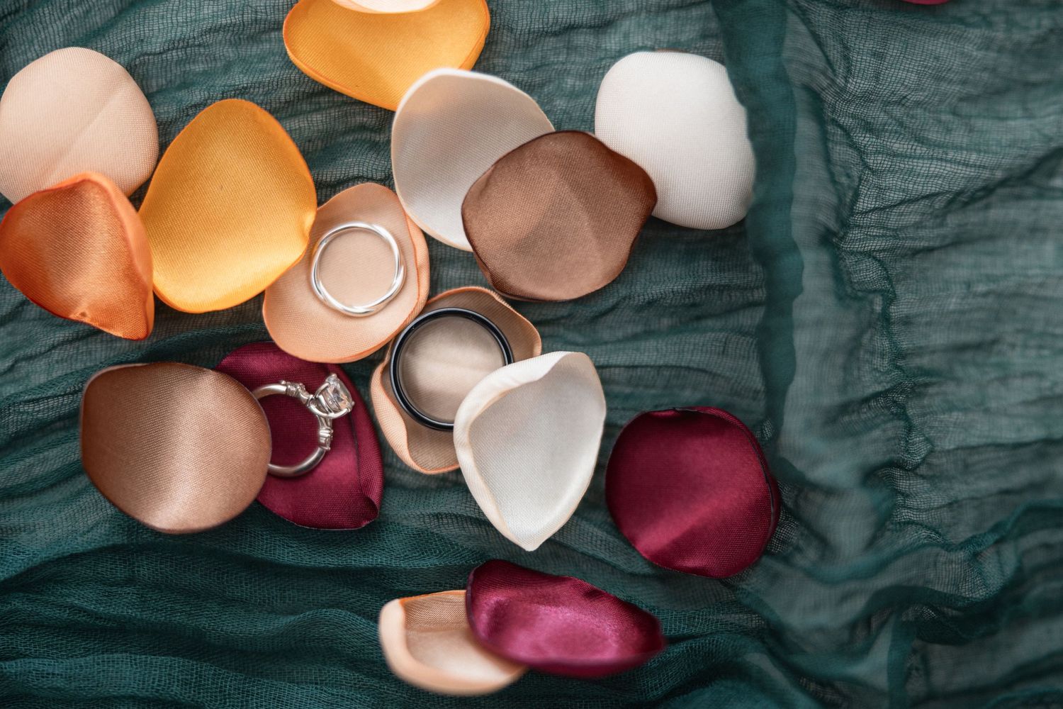 Collection of scattered rose gold, burgundy, and white silk rose petals on teal fabric.