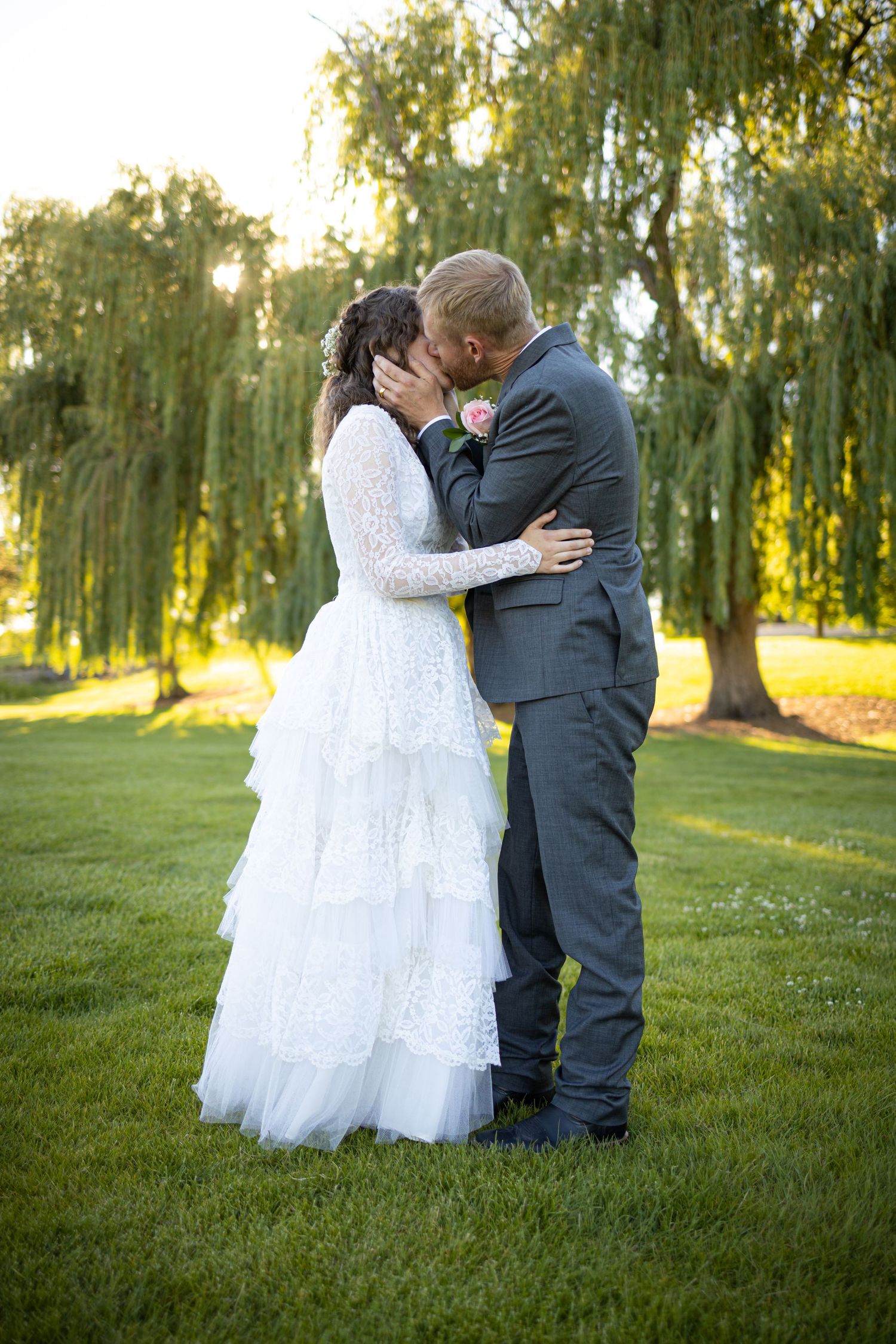 A romantic wedding couple sharing a kiss under weeping willow trees at sunset in a garden setting.
