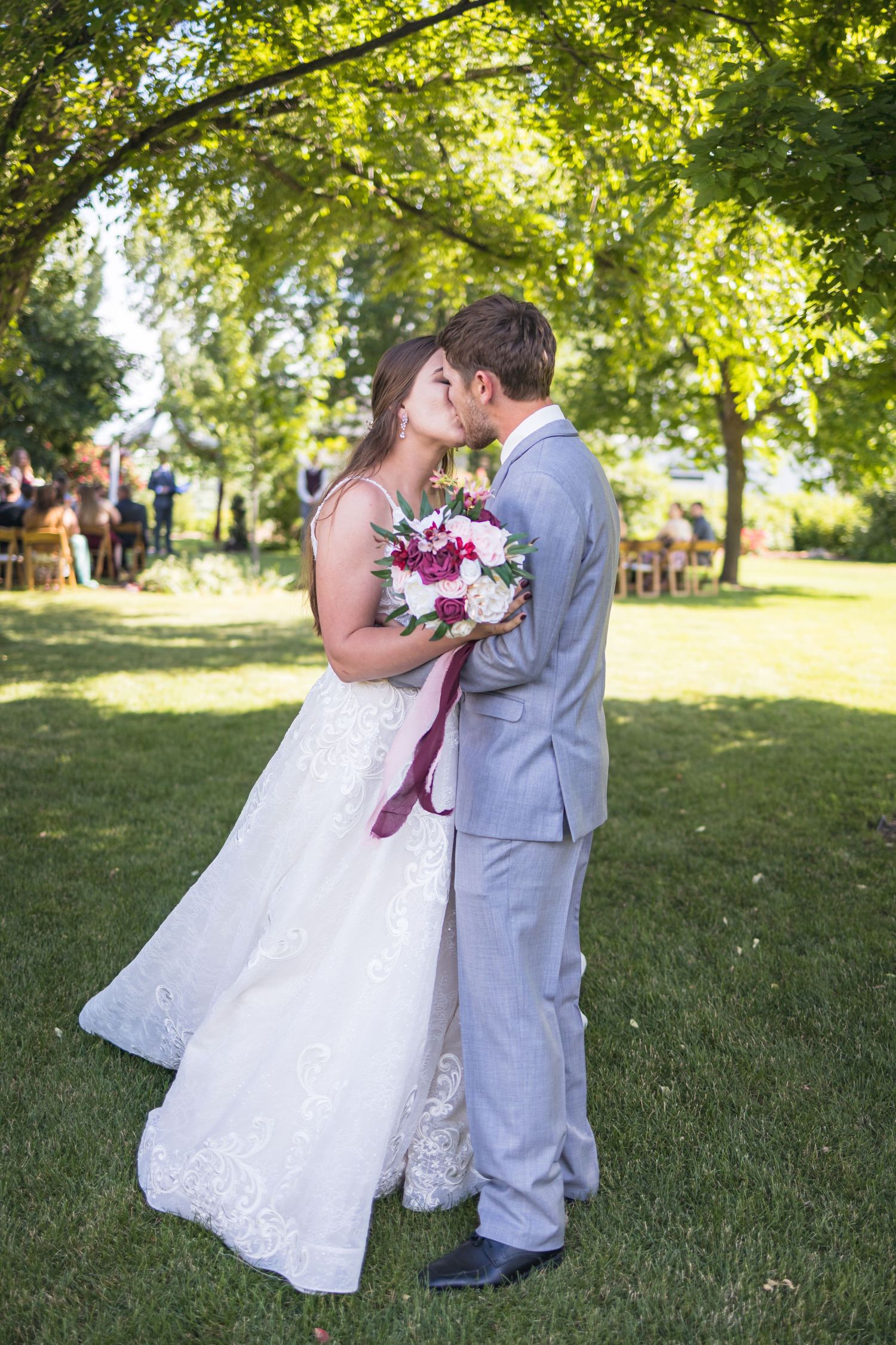 Bride and groom embracing during outdoor wedding photoshoot under green trees.