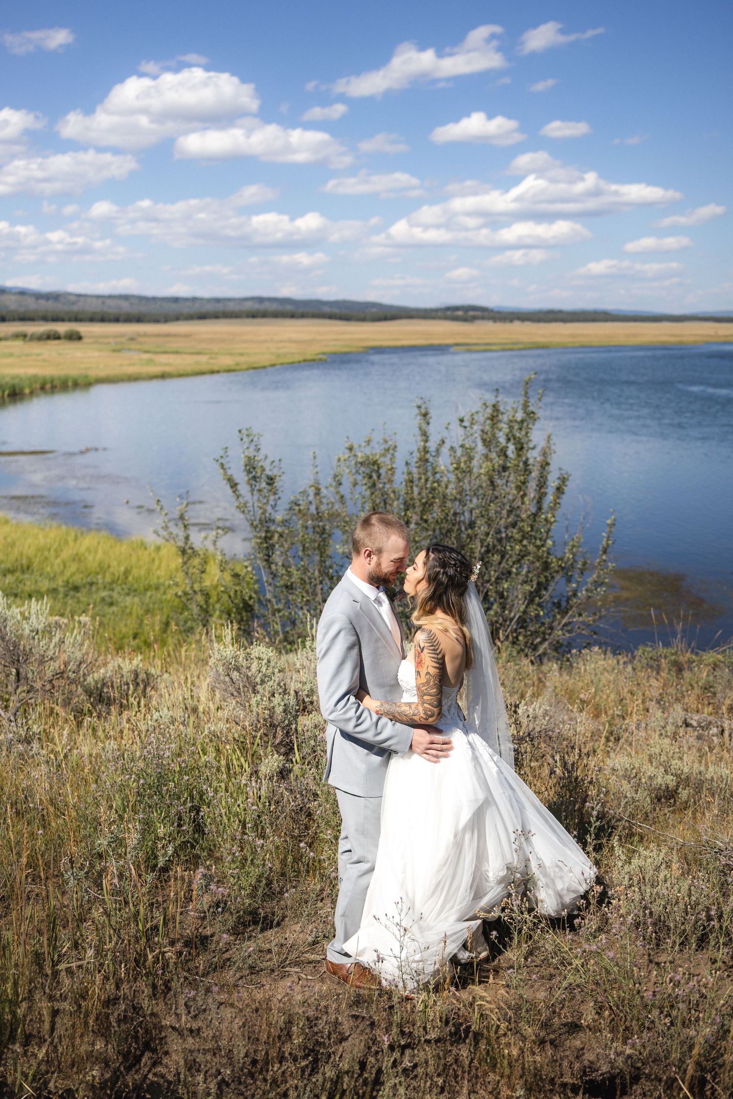 A romantic lakeside wedding photoshoot with mountains and blue skies in the background.