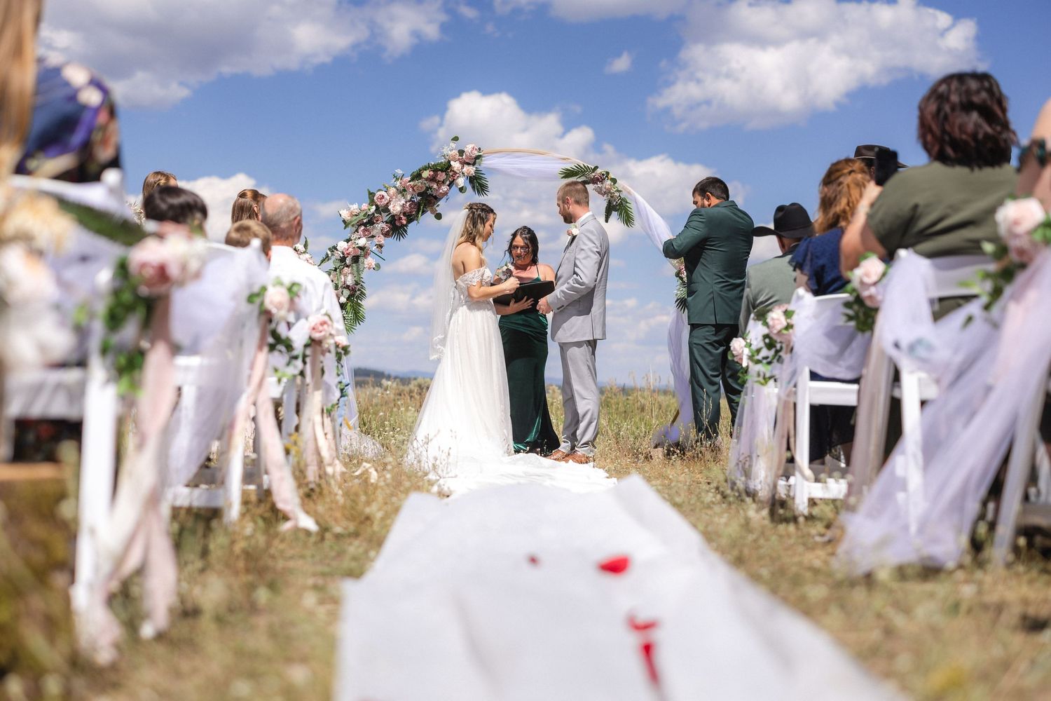 Outdoor wedding ceremony with white chairs and floral arch on a sunny mountain day.