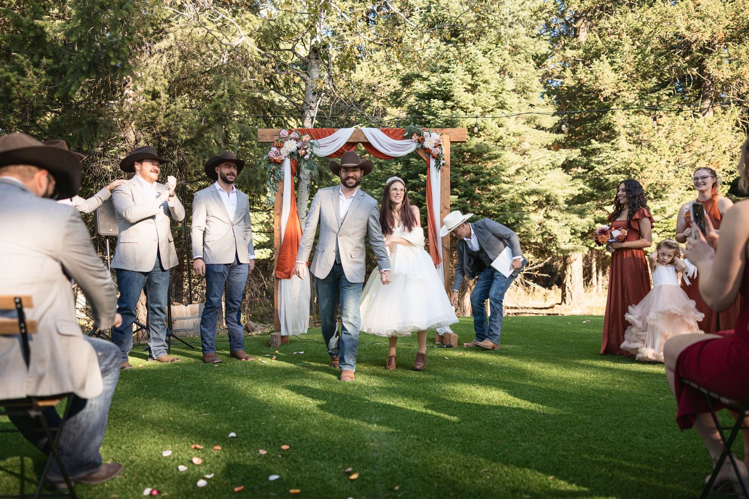 Newlyweds walking down aisle after ceremony under decorated wooden arch with guests celebrating.