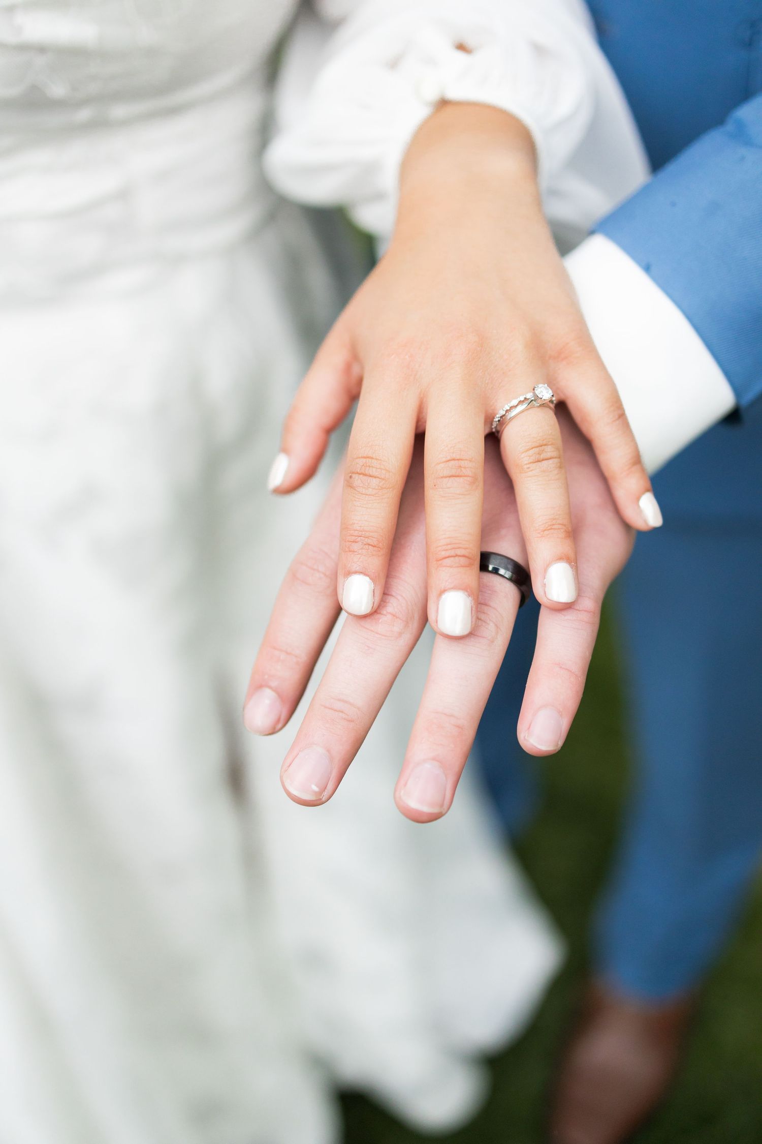 Close-up of newlywed couple's hands showing wedding rings against white dress and blue suit.