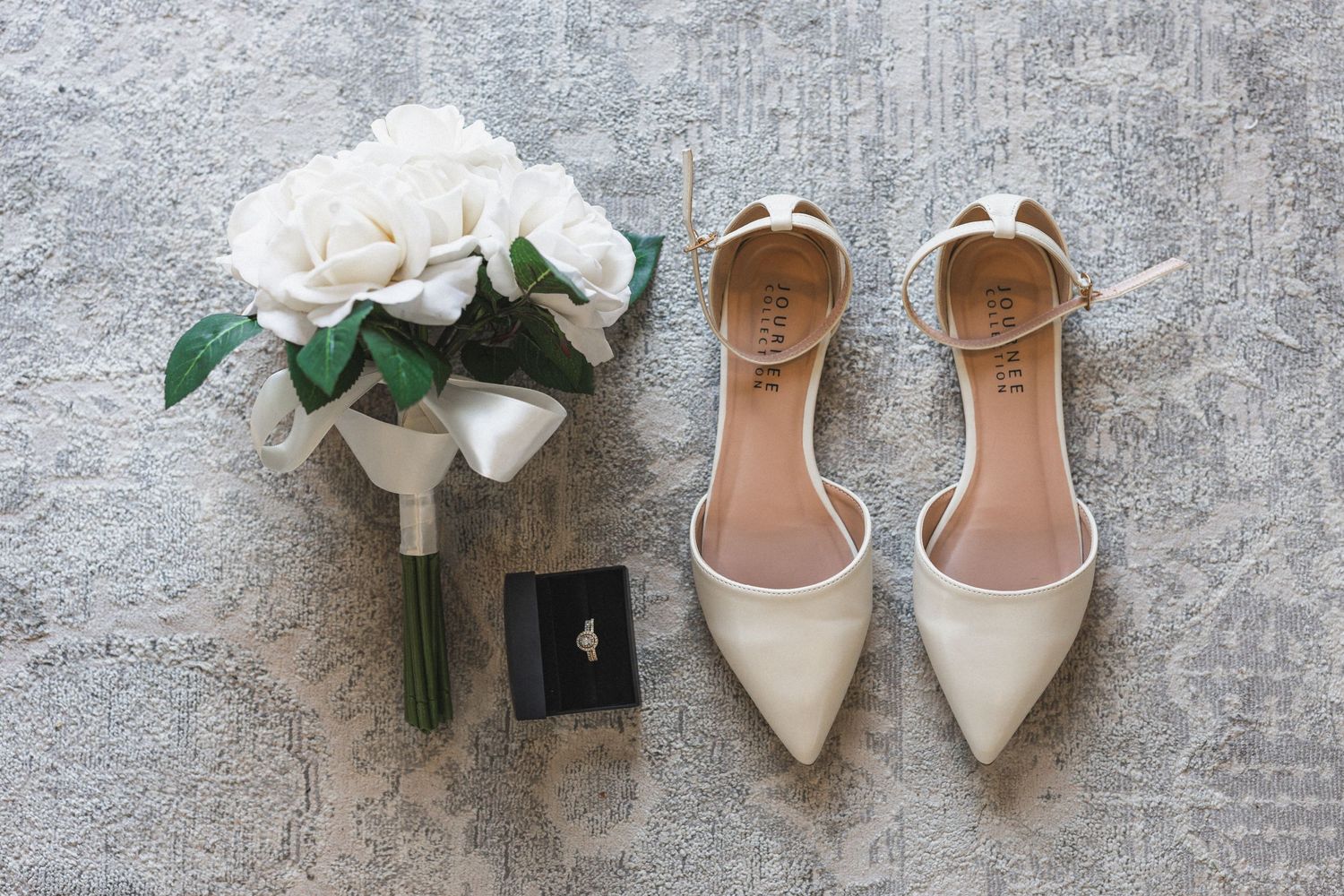 White bridal flats displayed with white rose bouquet and black ring box on stone floor.