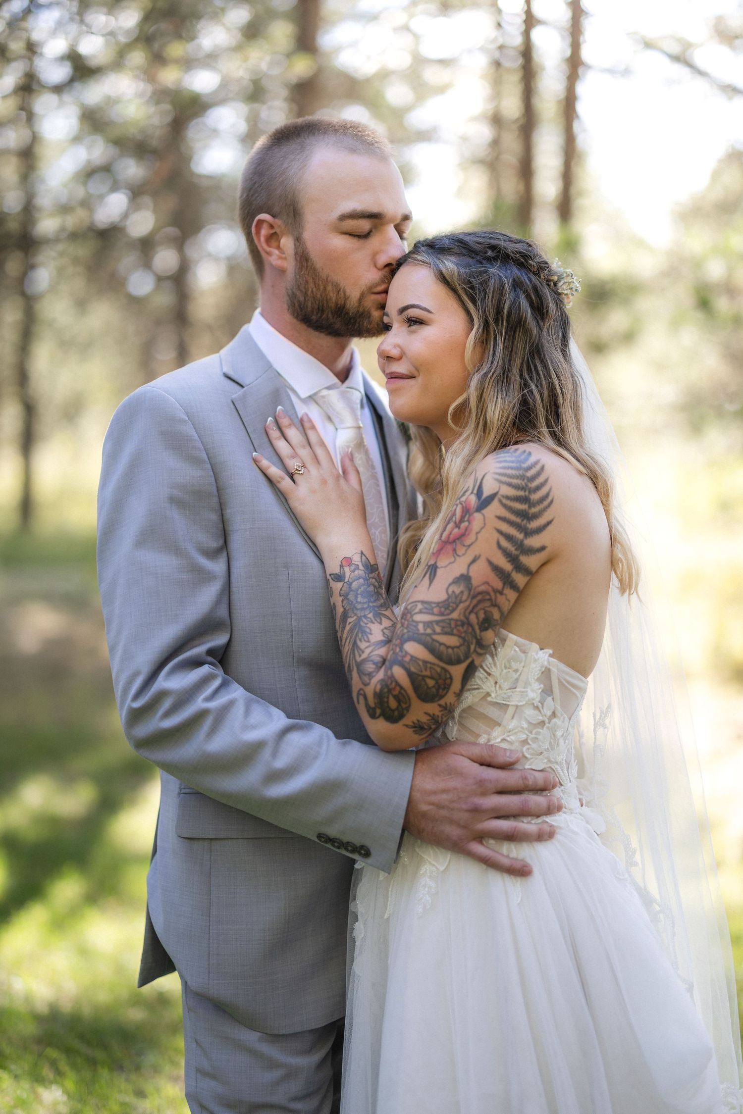 Newlywed couple in wedding attire embracing in a sun-dappled forest setting.