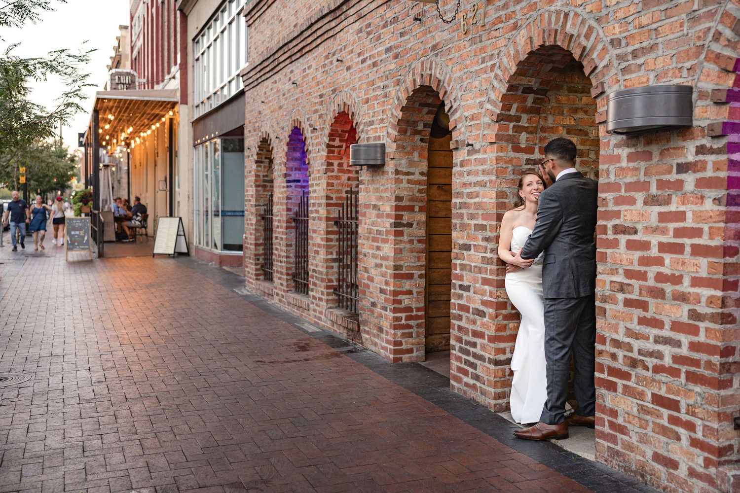 A couple embraces in a brick archway along a historic street at dusk.