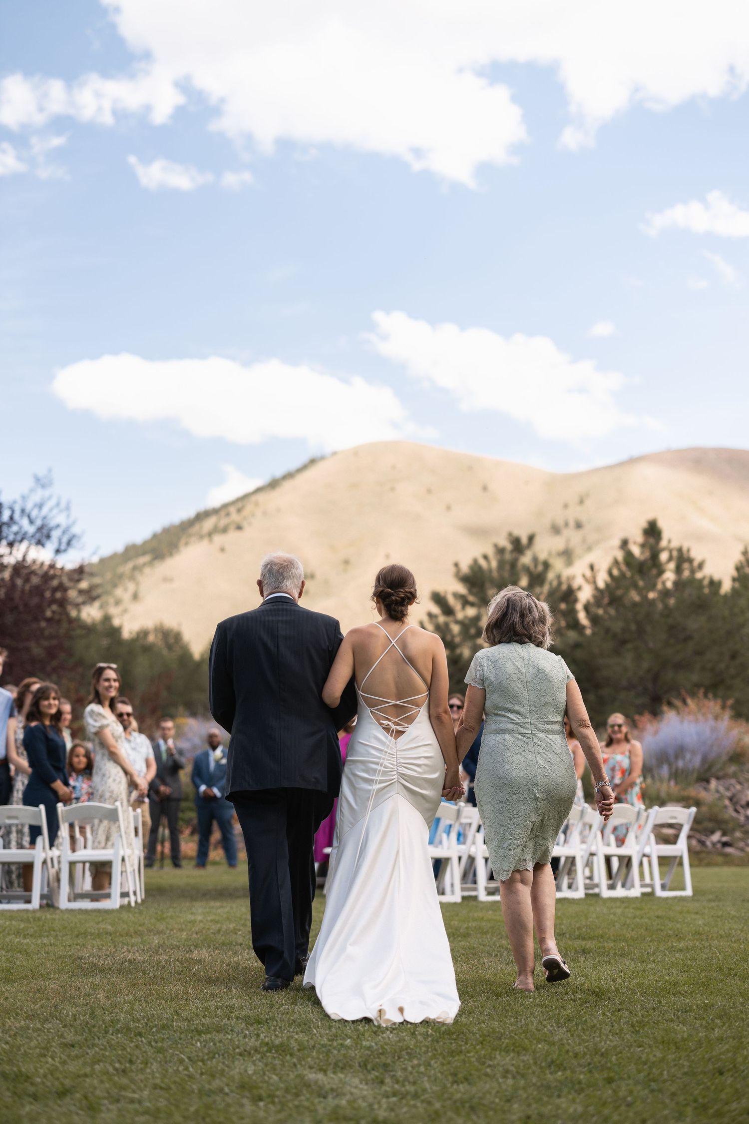 Wedding procession walks across green lawn with mountain views in background.
