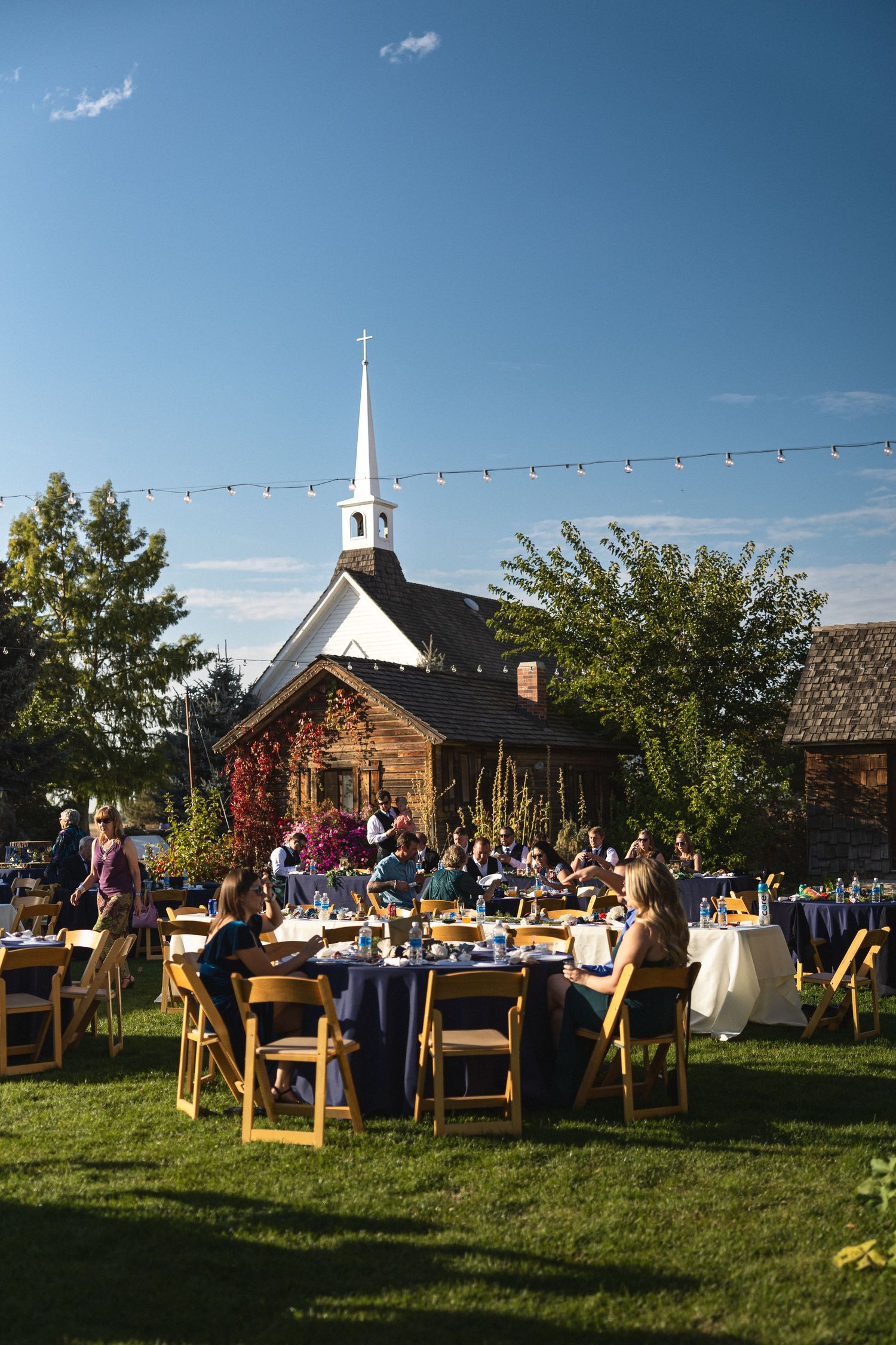 Outdoor wedding reception with wooden tables near charming chapel under string lights.