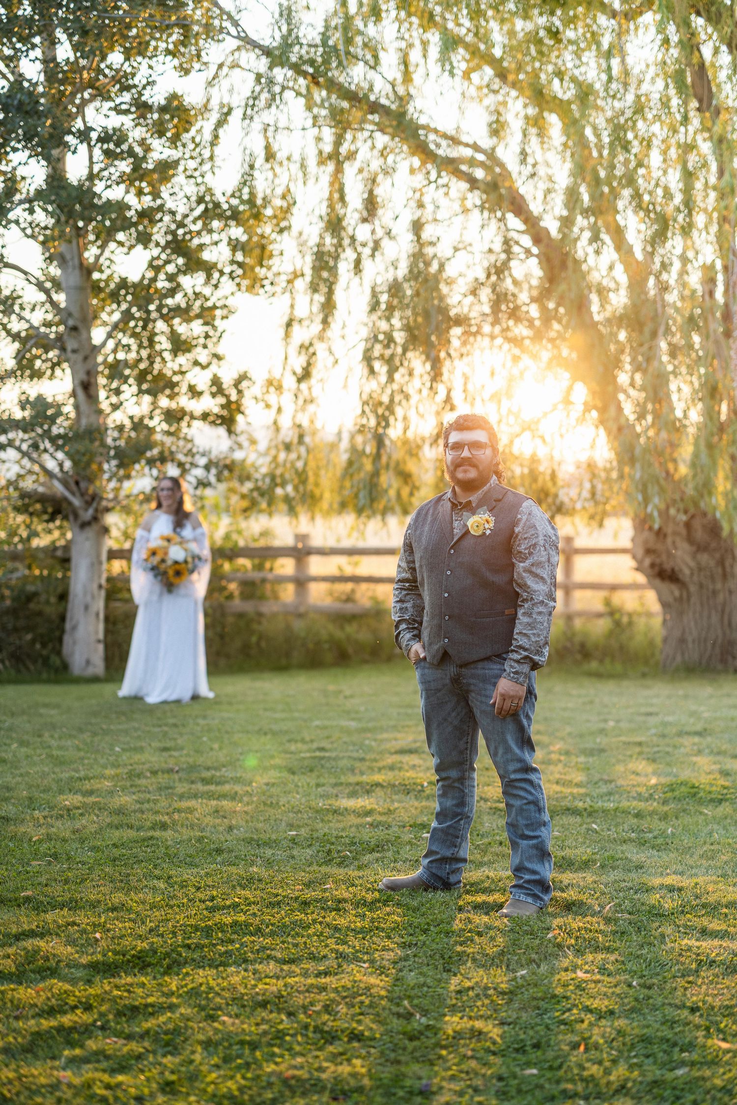 Wedding couple standing in grassy field with willow trees and golden sunset light.