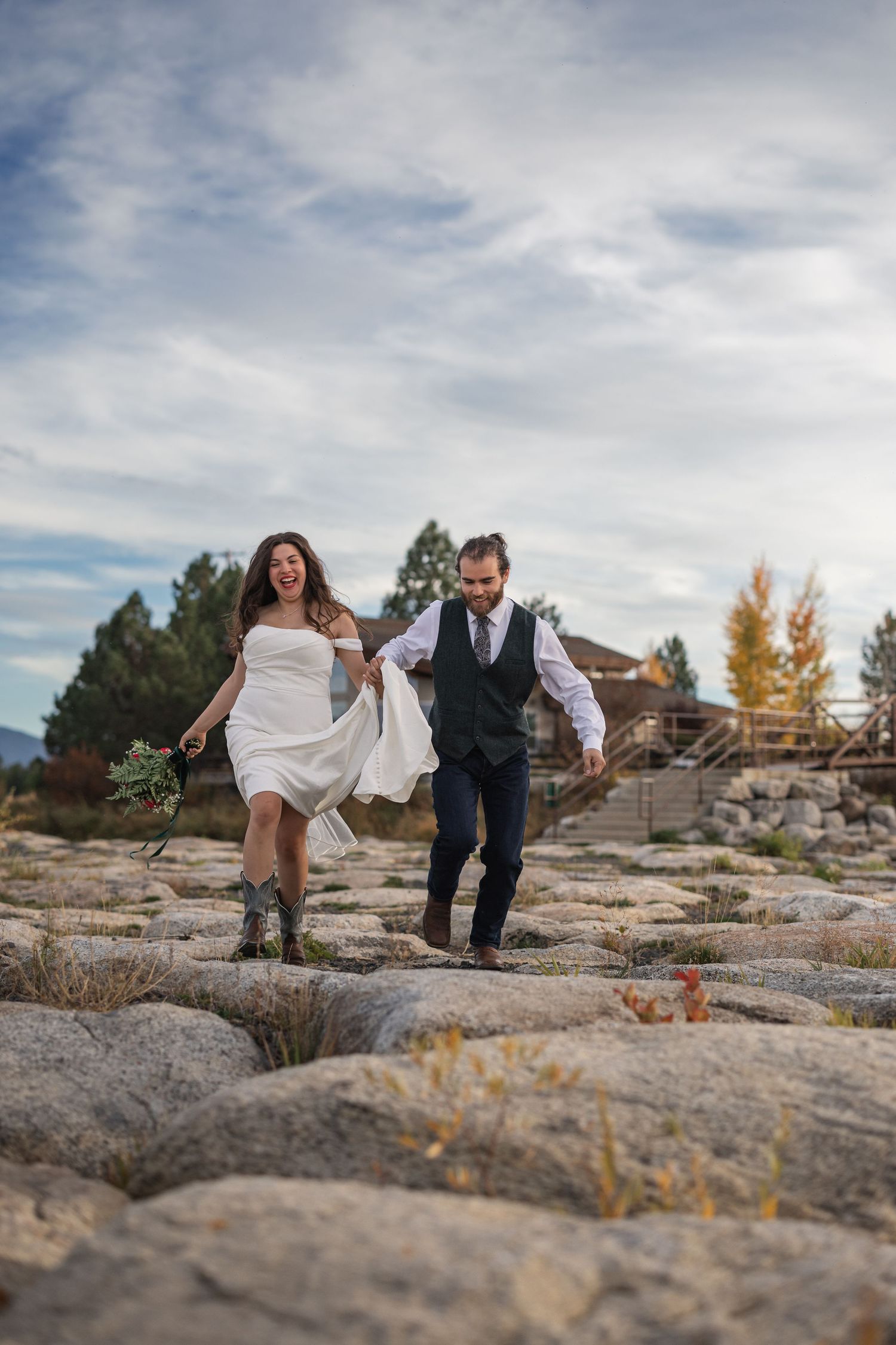 A couple running and laughing together on rocky terrain with a cloudy sky backdrop in wedding attire.