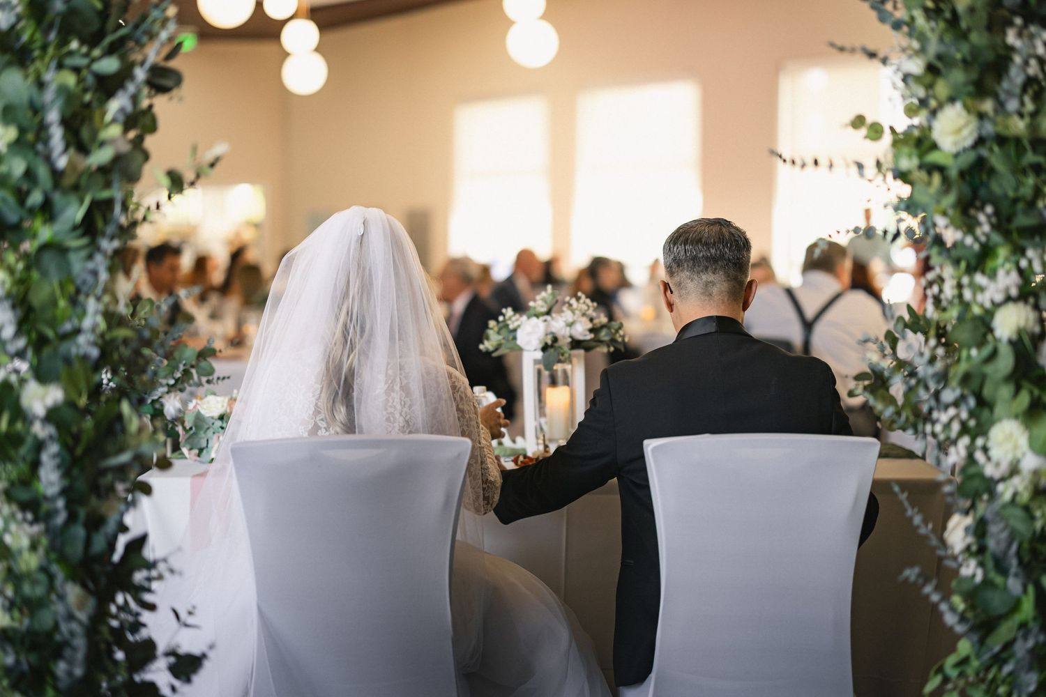 A couple seated at a wedding ceremony surrounded by greenery and hanging lights.