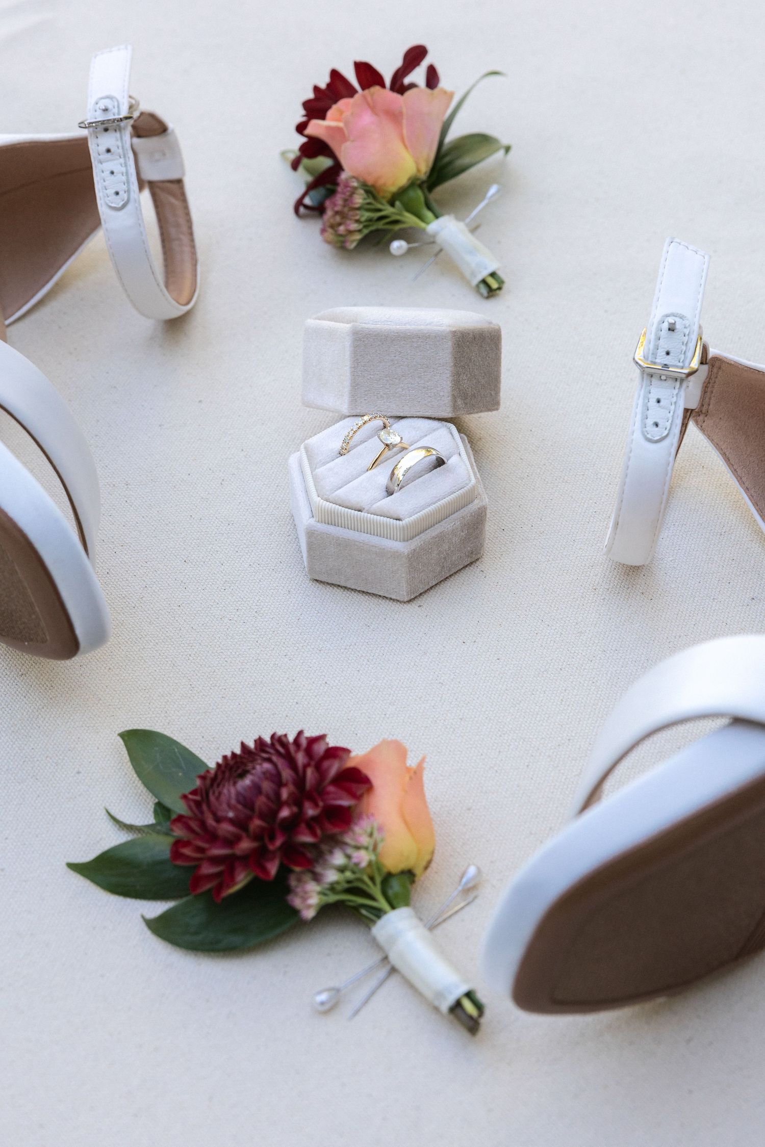 Wedding accessories including floral boutonnieres, heels, and a hexagonal ring box on light surface.