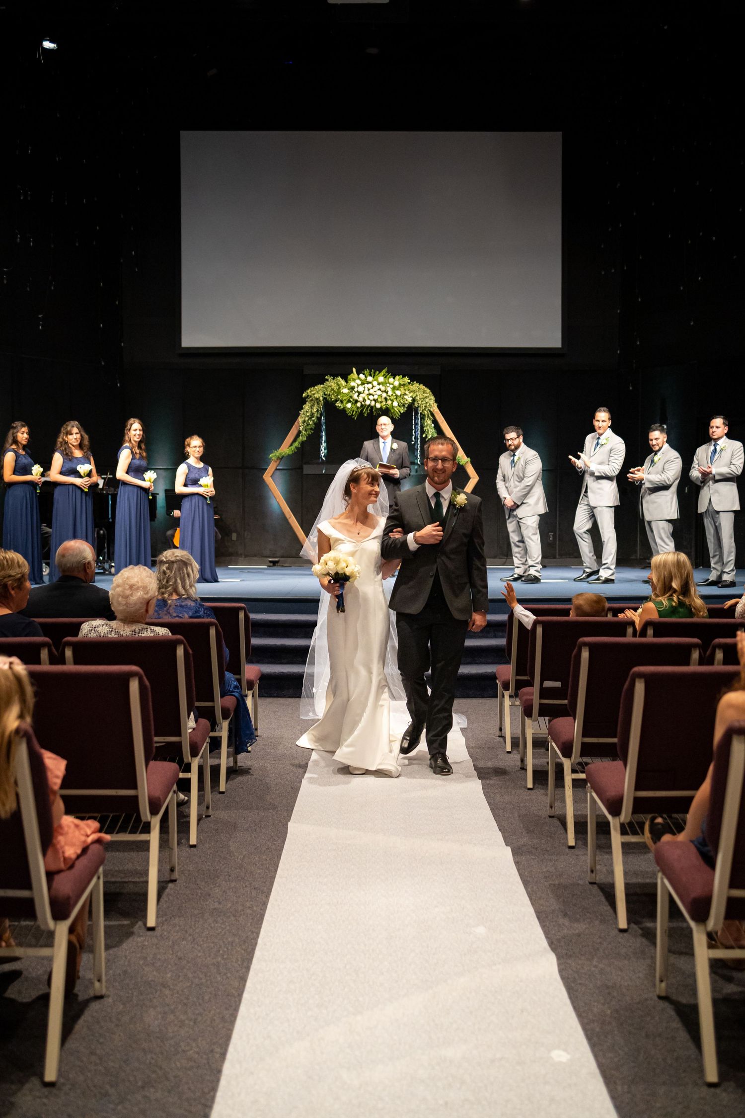 Newlyweds exit down white aisle runner with bridal party in navy and gray on either side.