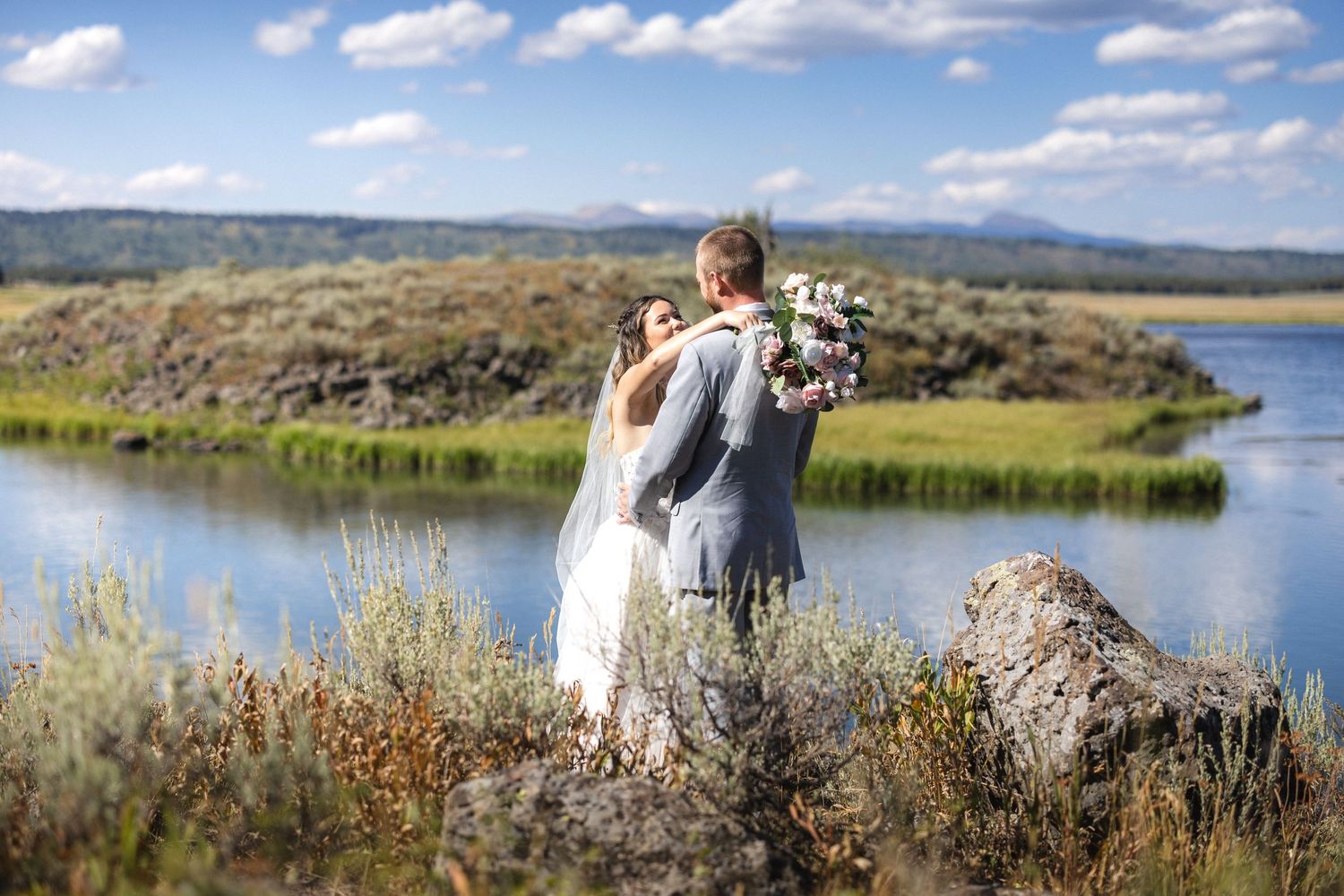 A wedding couple embraces on a rocky overlook with a scenic lake and mountains in the background.