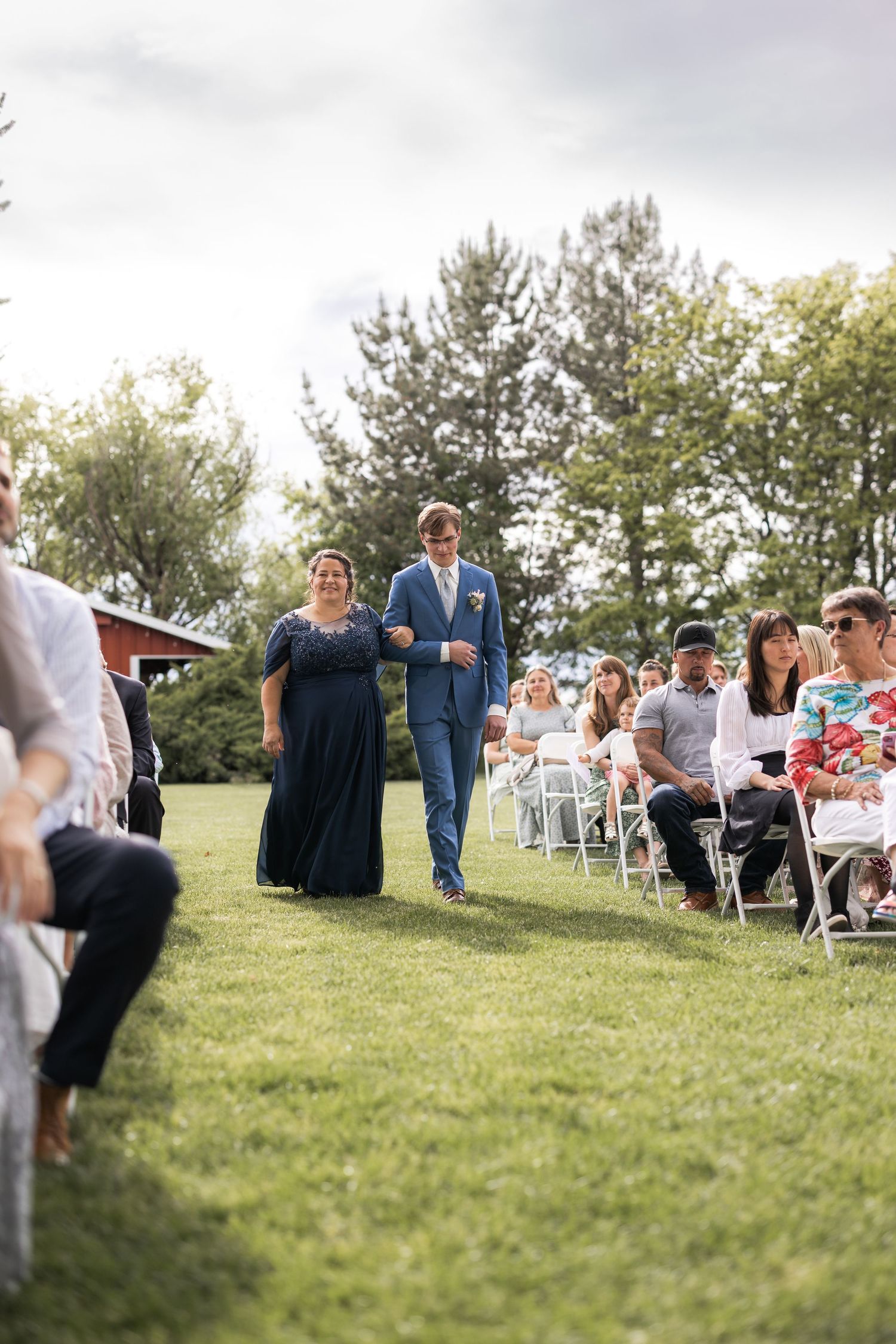 Wedding processional on a grassy lawn with guests seated in white chairs and trees in background.
