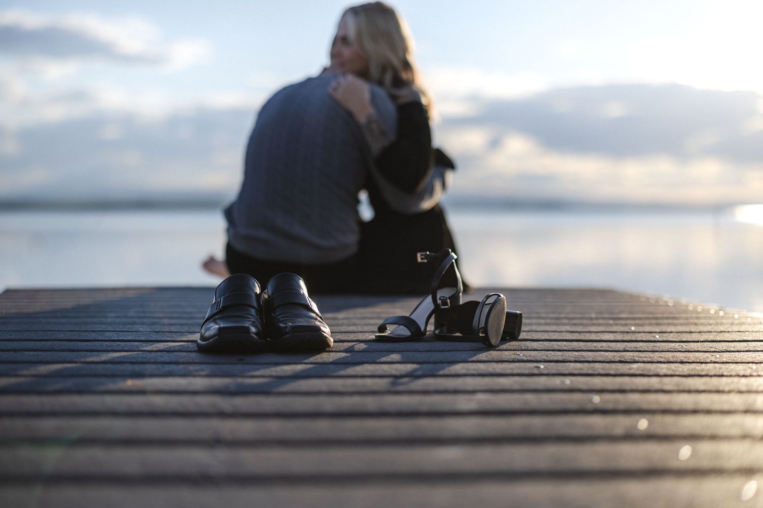 Person sitting alone on wooden dock at sunset, photographed from behind with shallow depth of field.