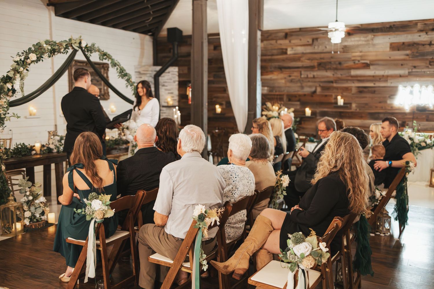 Wedding guests seated in rows at rustic indoor venue with wood walls and floral arch decorations.