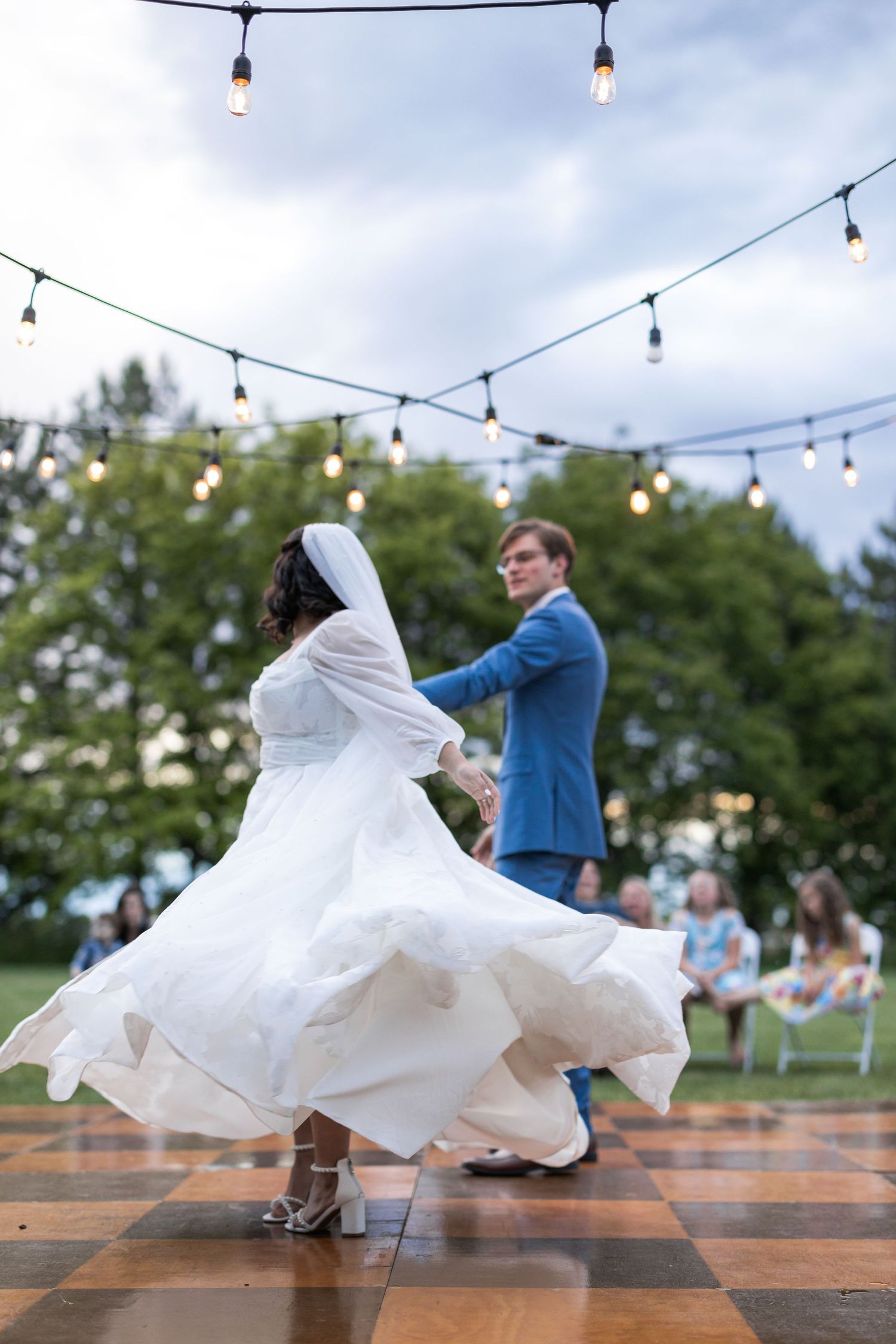 A couple dances on a wooden dance floor under string lights at an outdoor wedding reception.