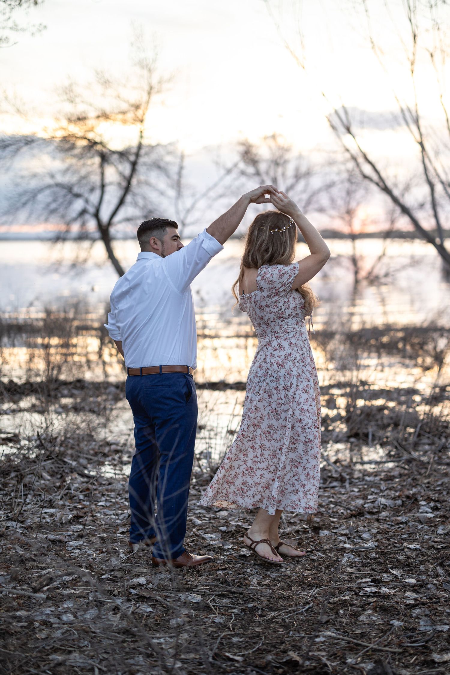 A couple dances together at sunset near a lake with bare winter trees in the background.