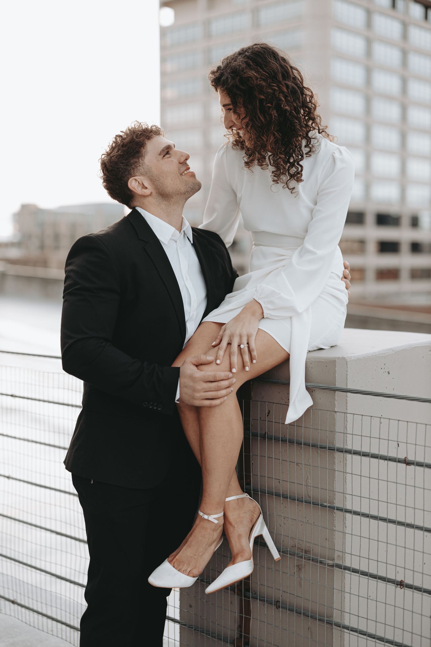 A romantic couple shares an intimate moment on a ledge, with urban architecture in the background.
