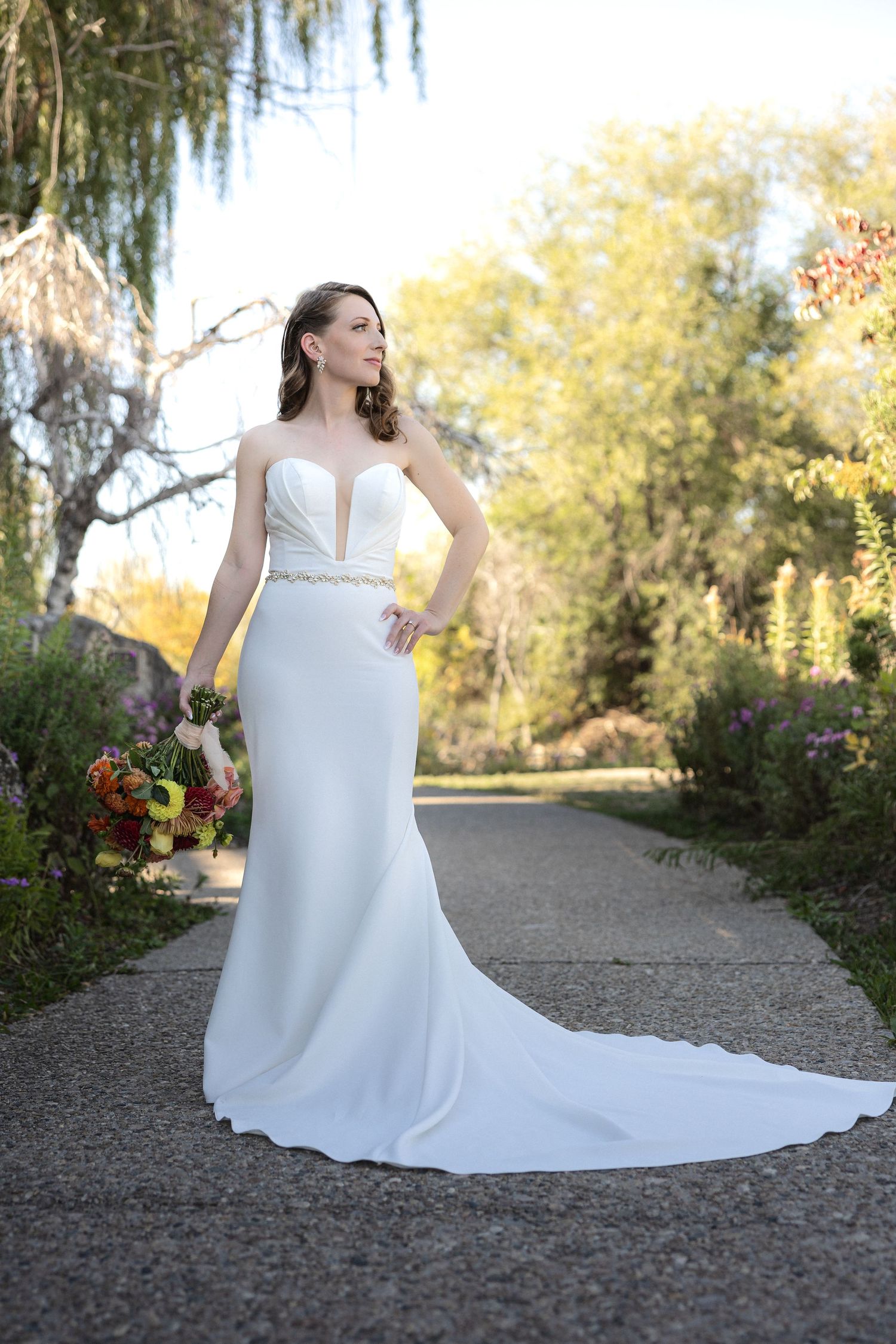 Bride in elegant white fitted wedding gown poses on garden path during autumn.