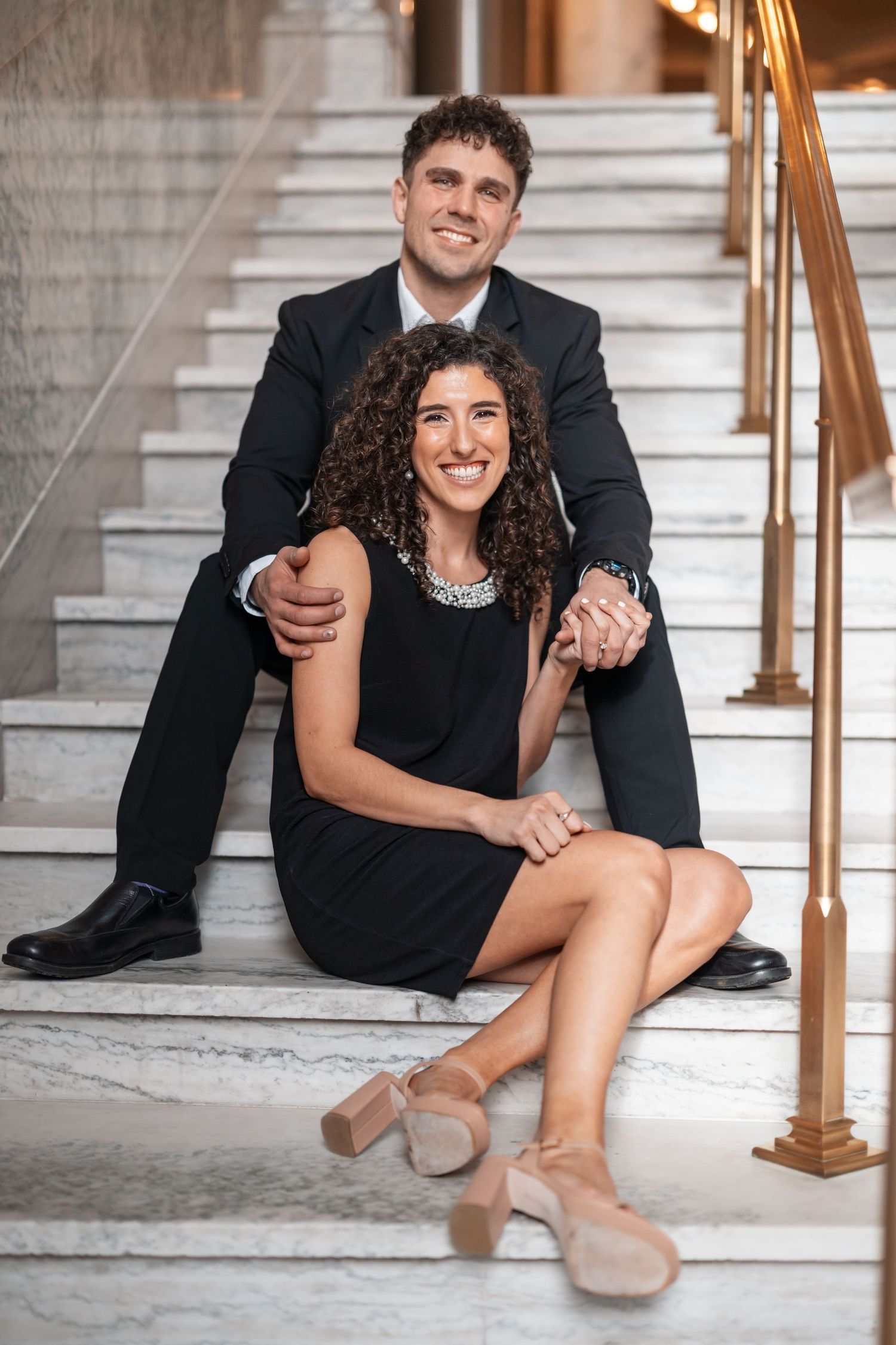 Couple in formal black attire pose intimately on elegant marble staircase with brass railing.