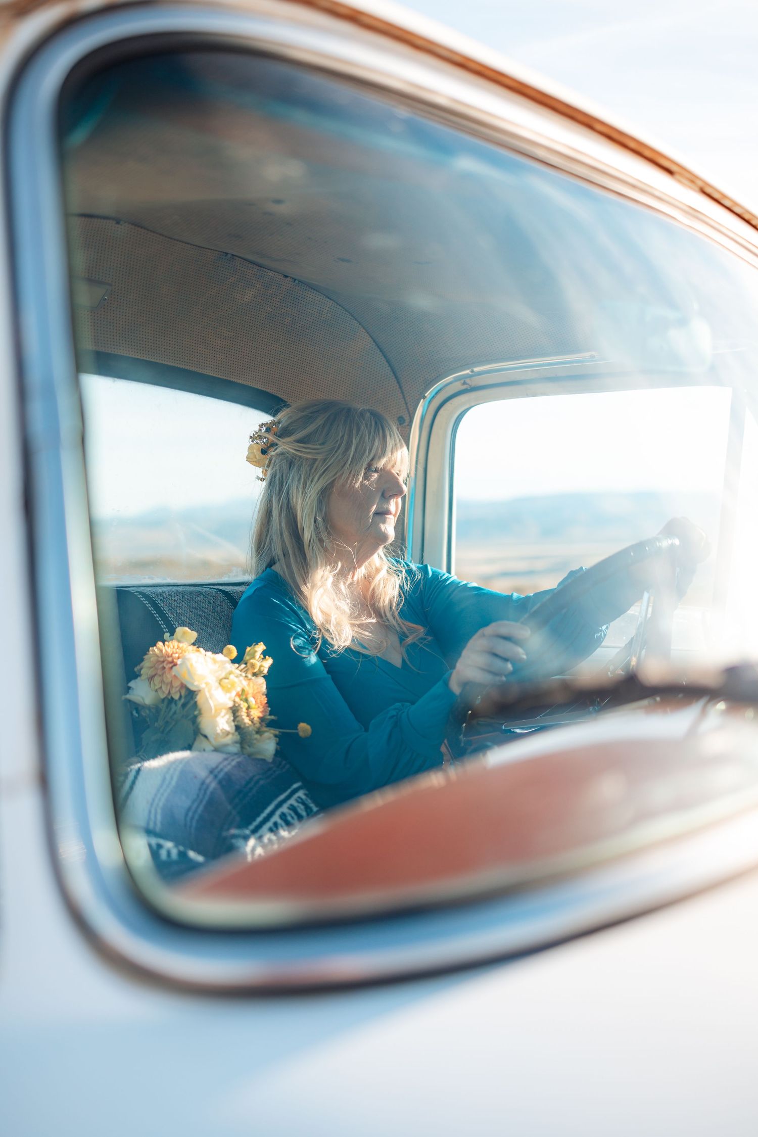 View through car window showing someone in a blue top driving with yellow flowers on the passenger seat.