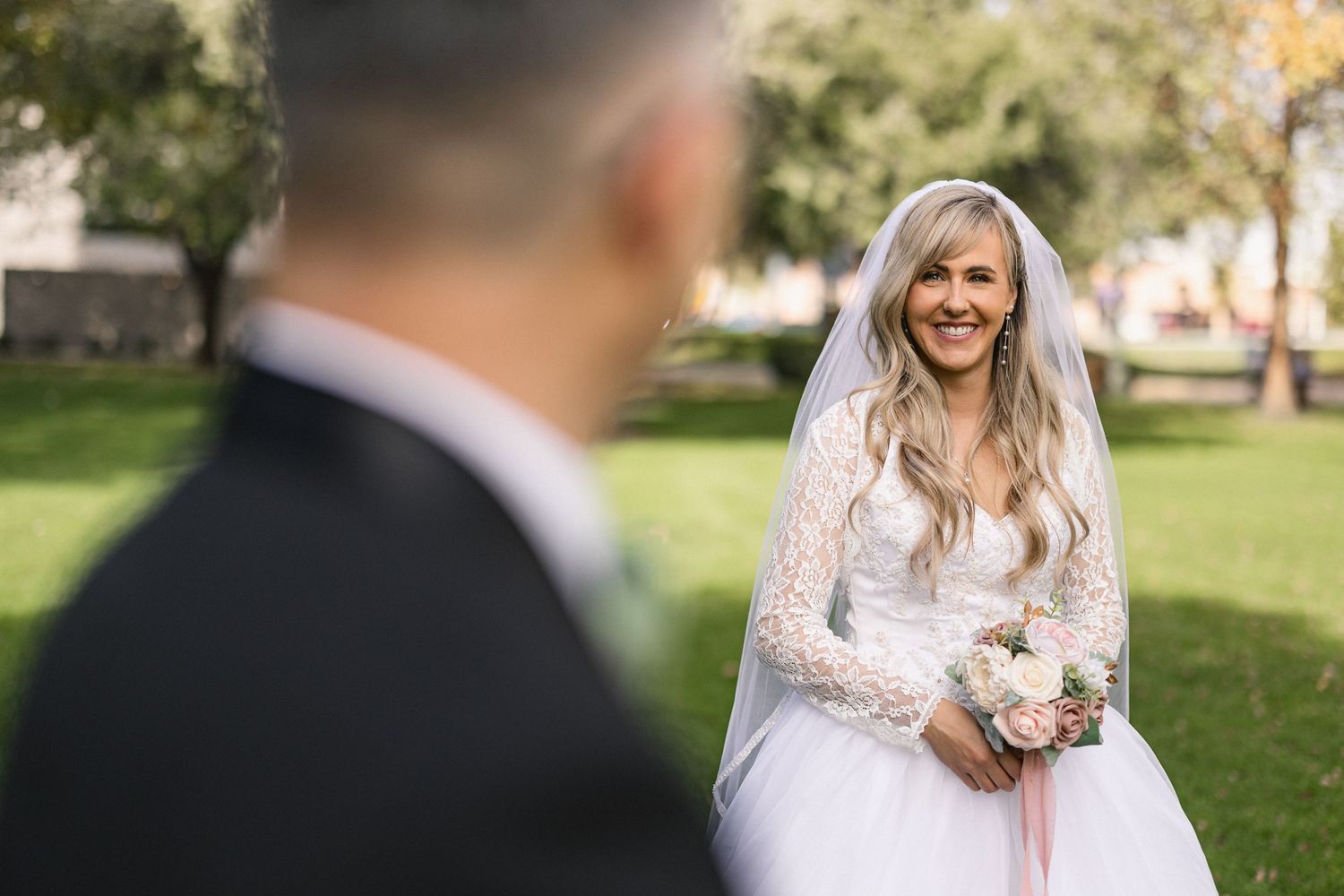Bride in white lace dress holding bouquet while groom looks on in garden setting.