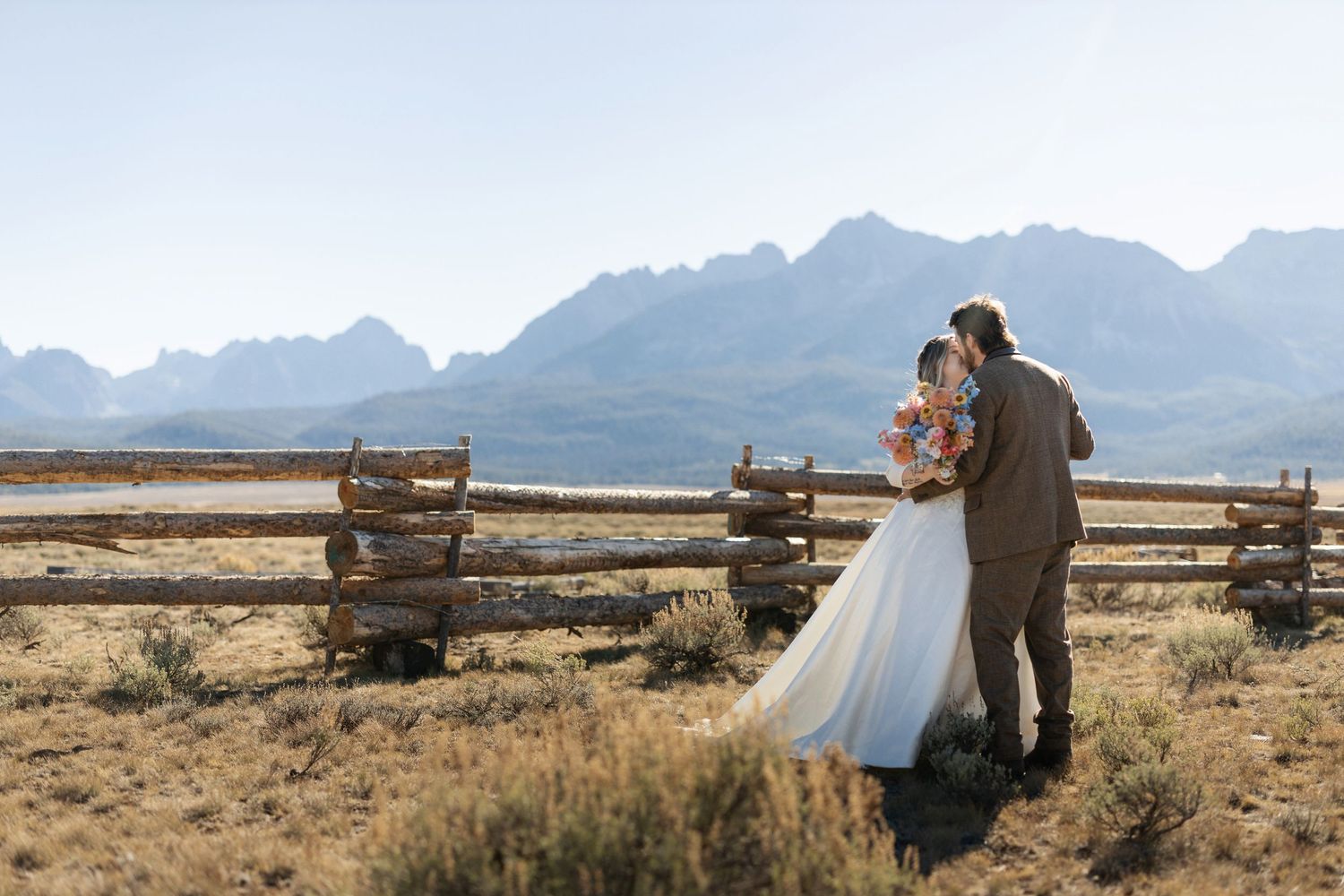Wedding couple walking hand in hand through open field with mountains and wooden fence in background.