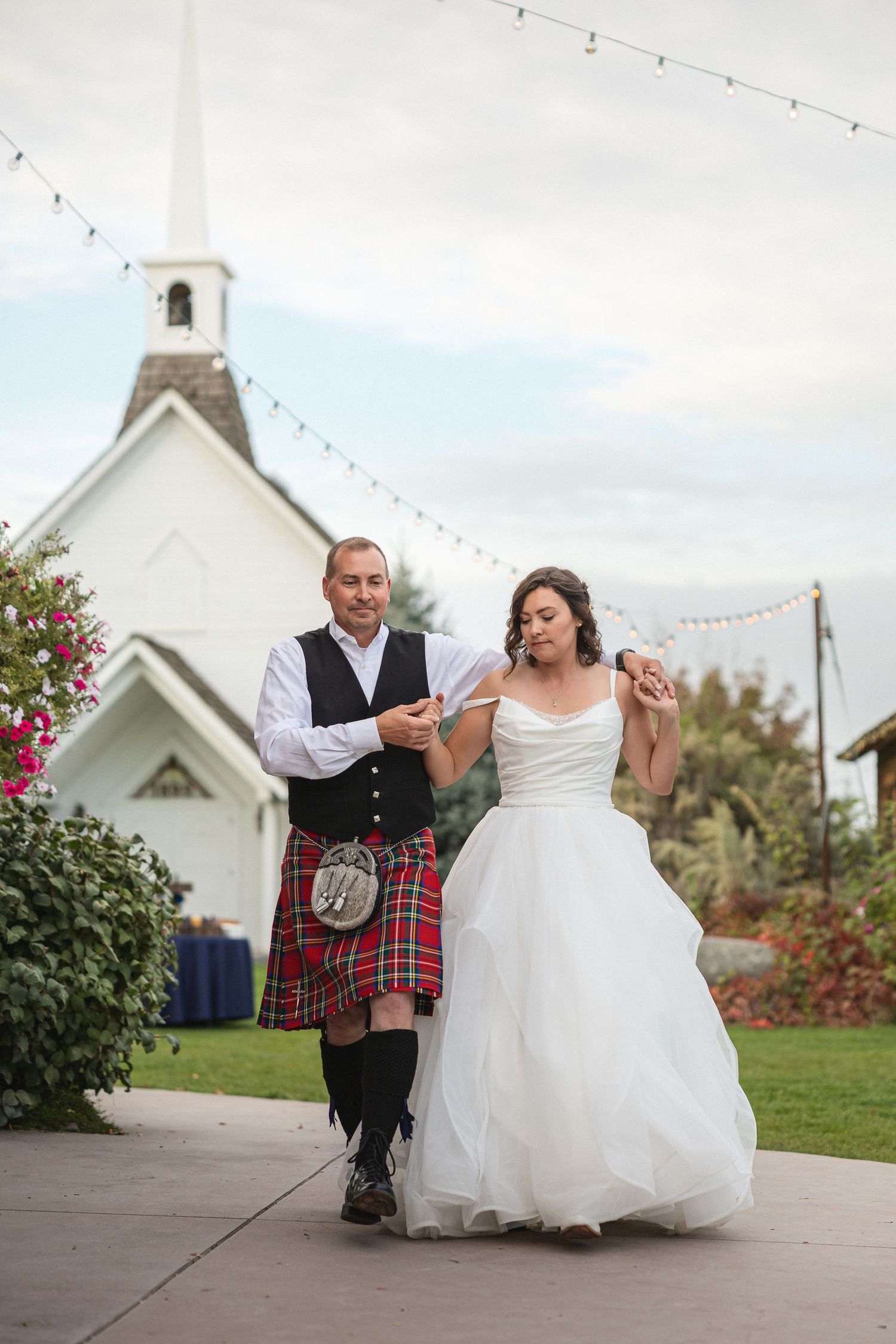 A couple dances on a patio with a white chapel and string lights creating a romantic outdoor setting.