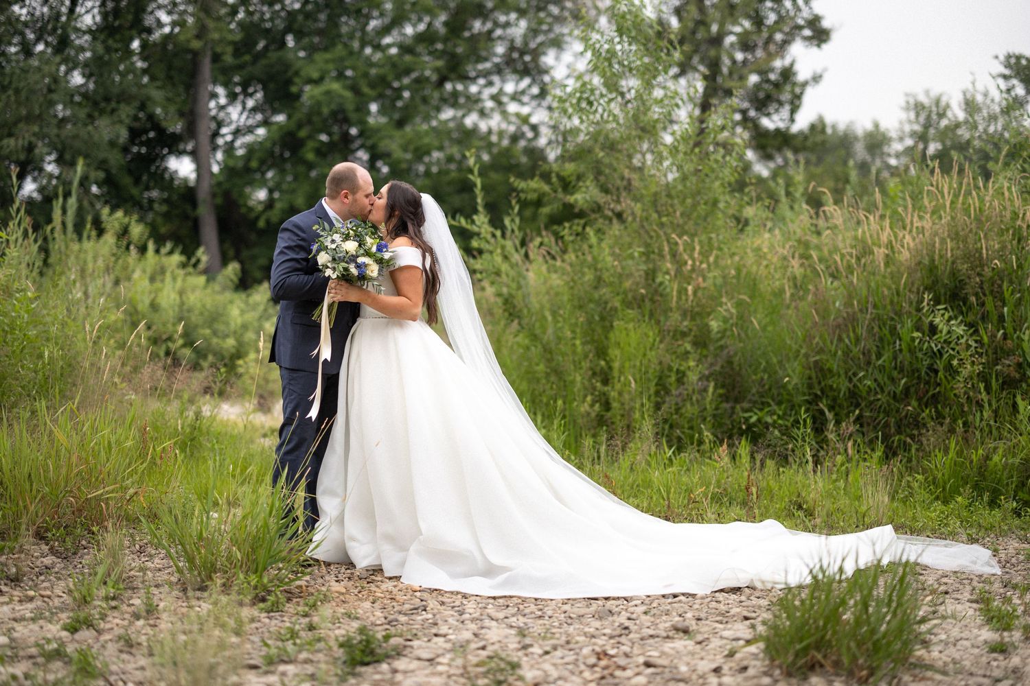 Wedding couple embraces on natural path surrounded by summer greenery.