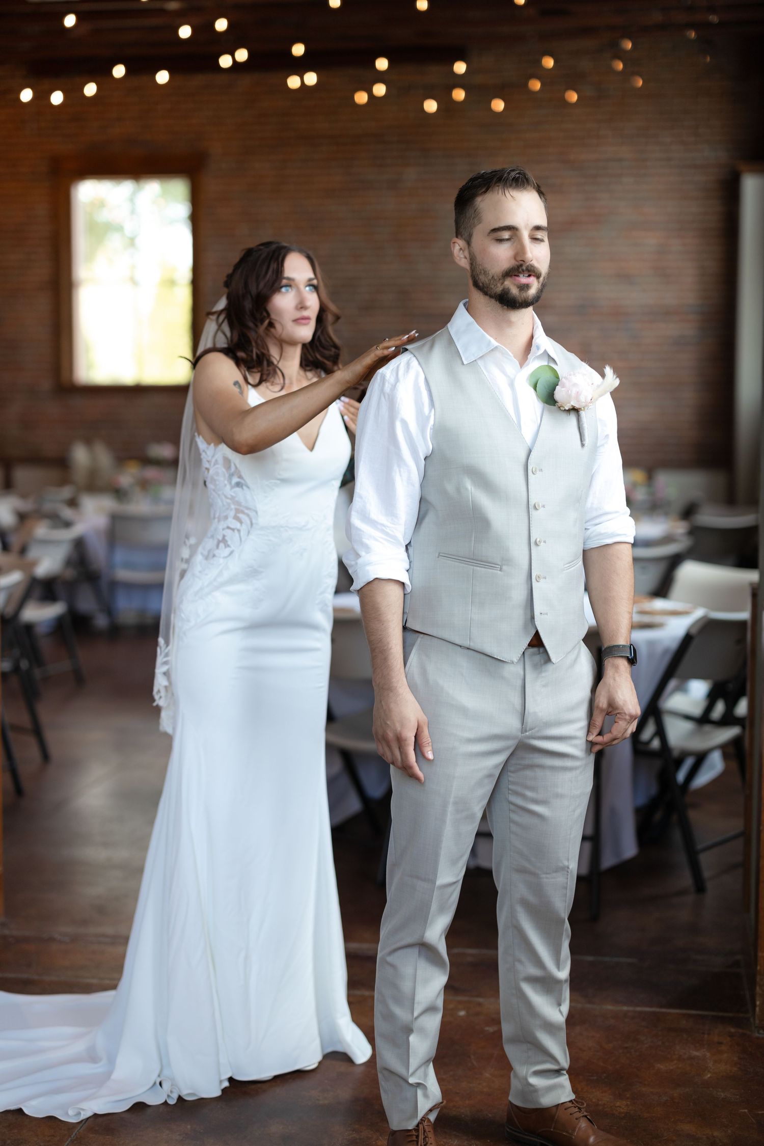 Wedding couple in a rustic brick venue with string lights, bride adjusting groom's boutonniere in elegant attire.