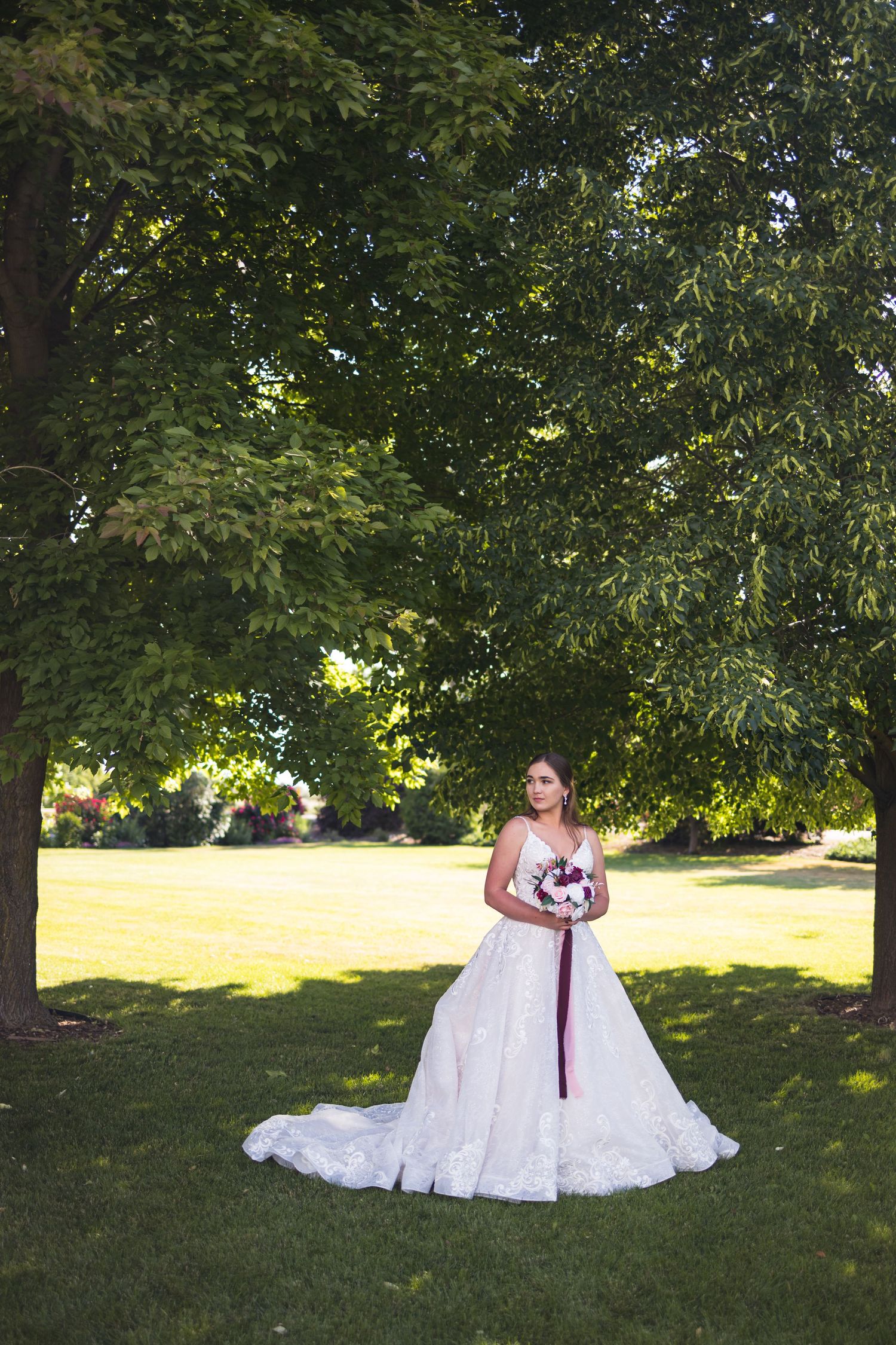 A bride in a white lace wedding dress poses under a shady tree on a sunny summer day.