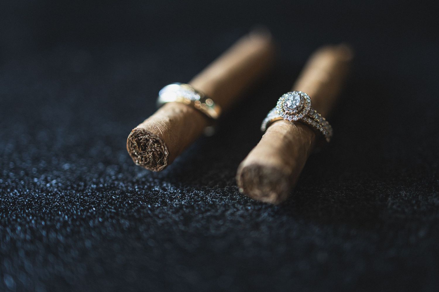 Wedding rings displayed artistically on two brown cigars with dark textured background.