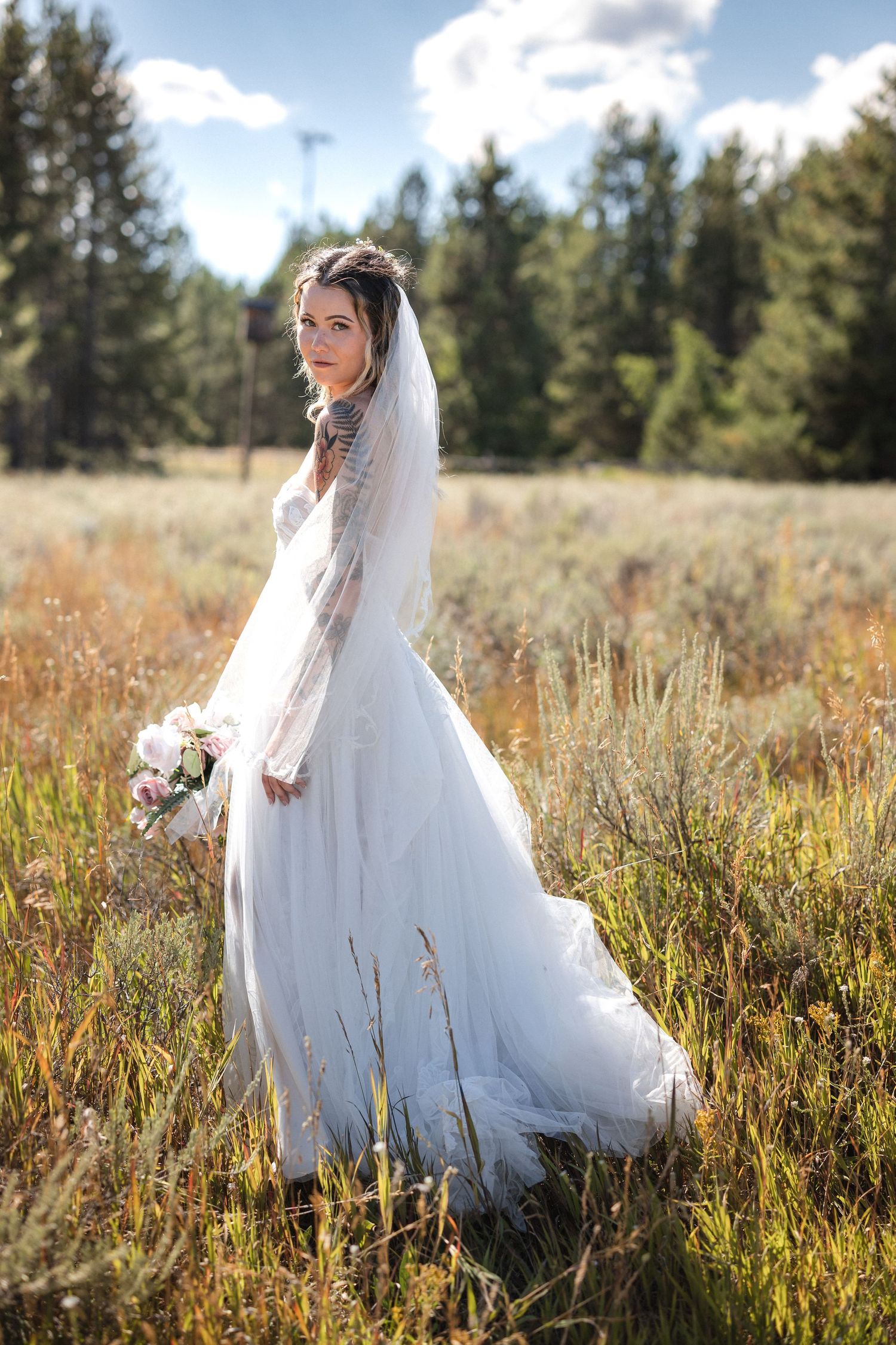A bride in a white gown stands in golden grassland holding a bouquet with evergreen forest backdrop.