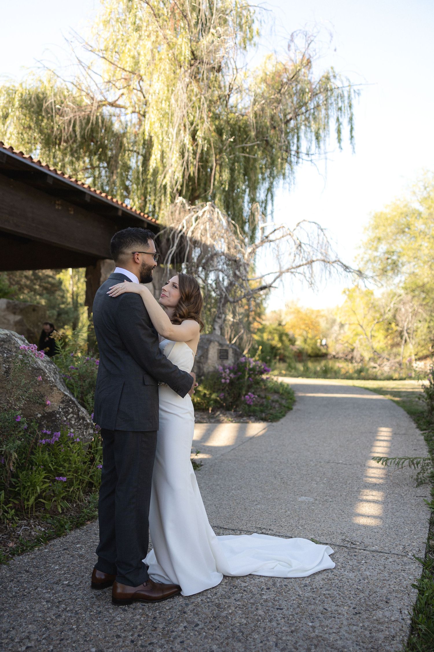 A couple embraces on a garden path beneath a weeping willow tree at their wedding.