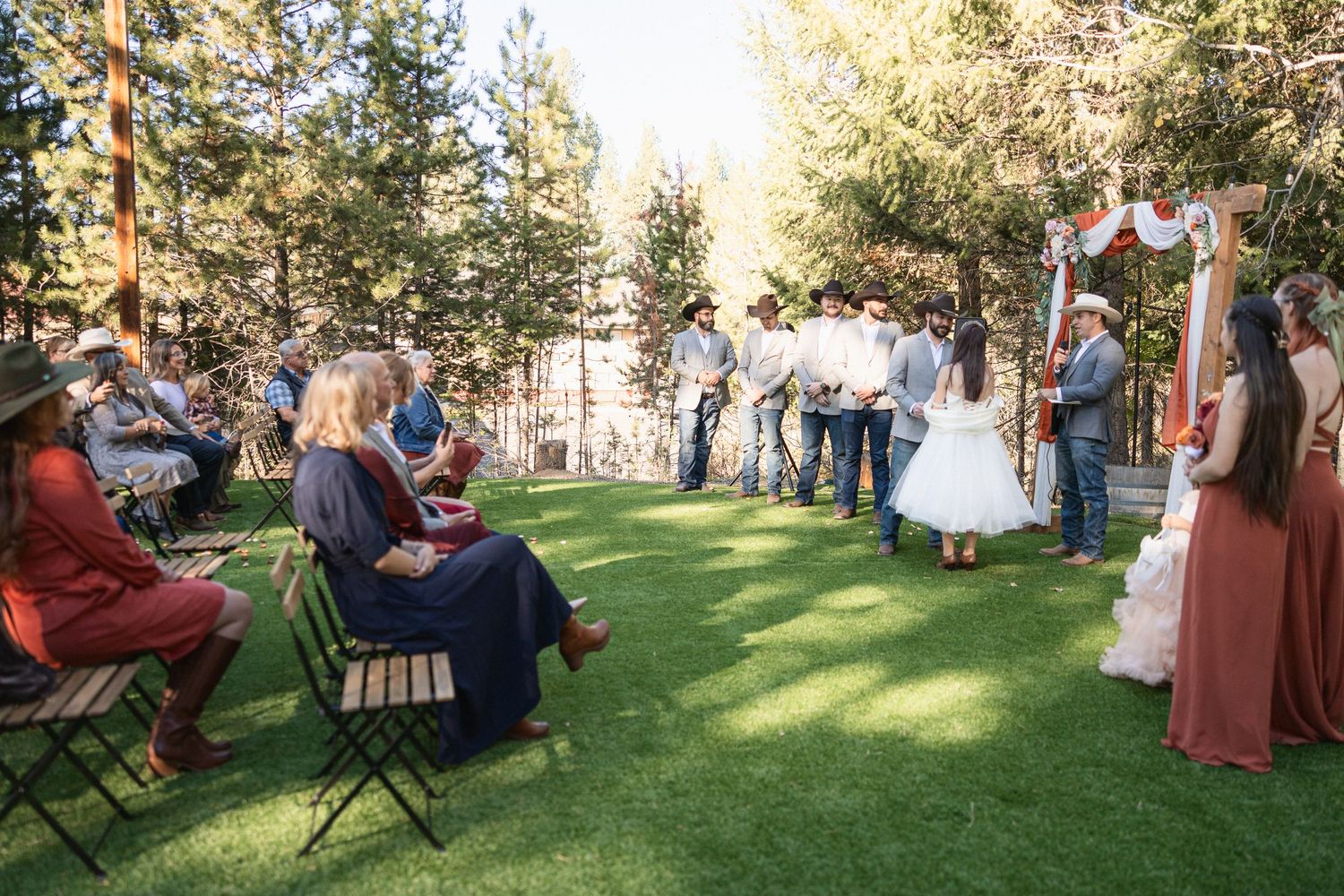 Outdoor wedding ceremony with guests seated on folding chairs on manicured lawn surrounded by pine trees.
