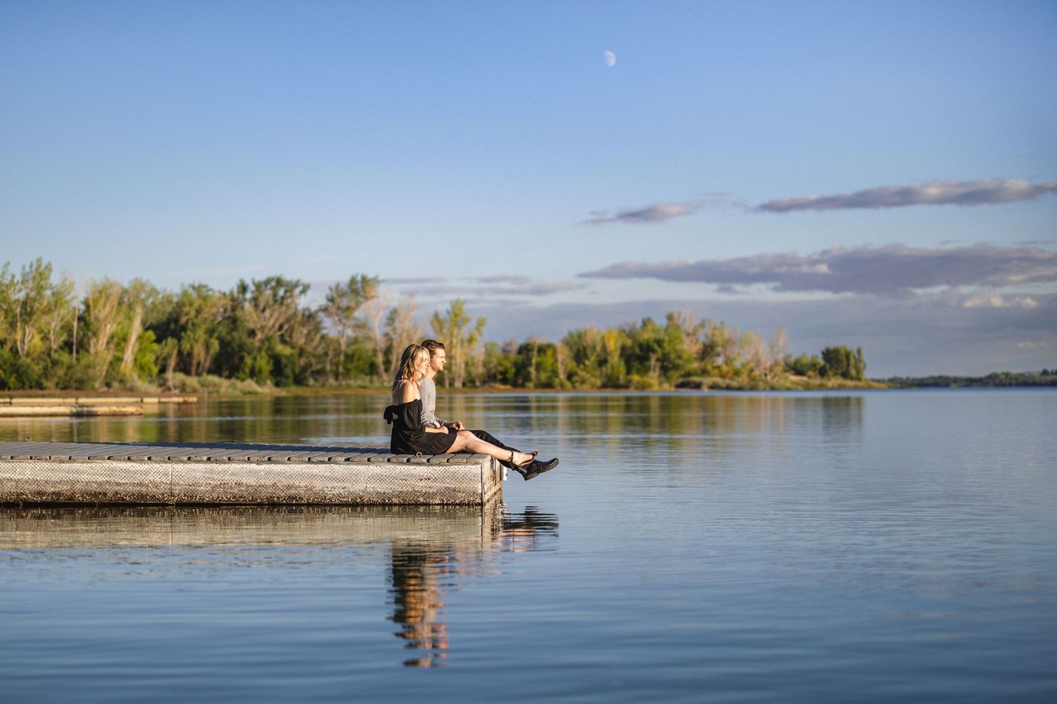 Person sitting on edge of dock with feet dangling over calm lake water.