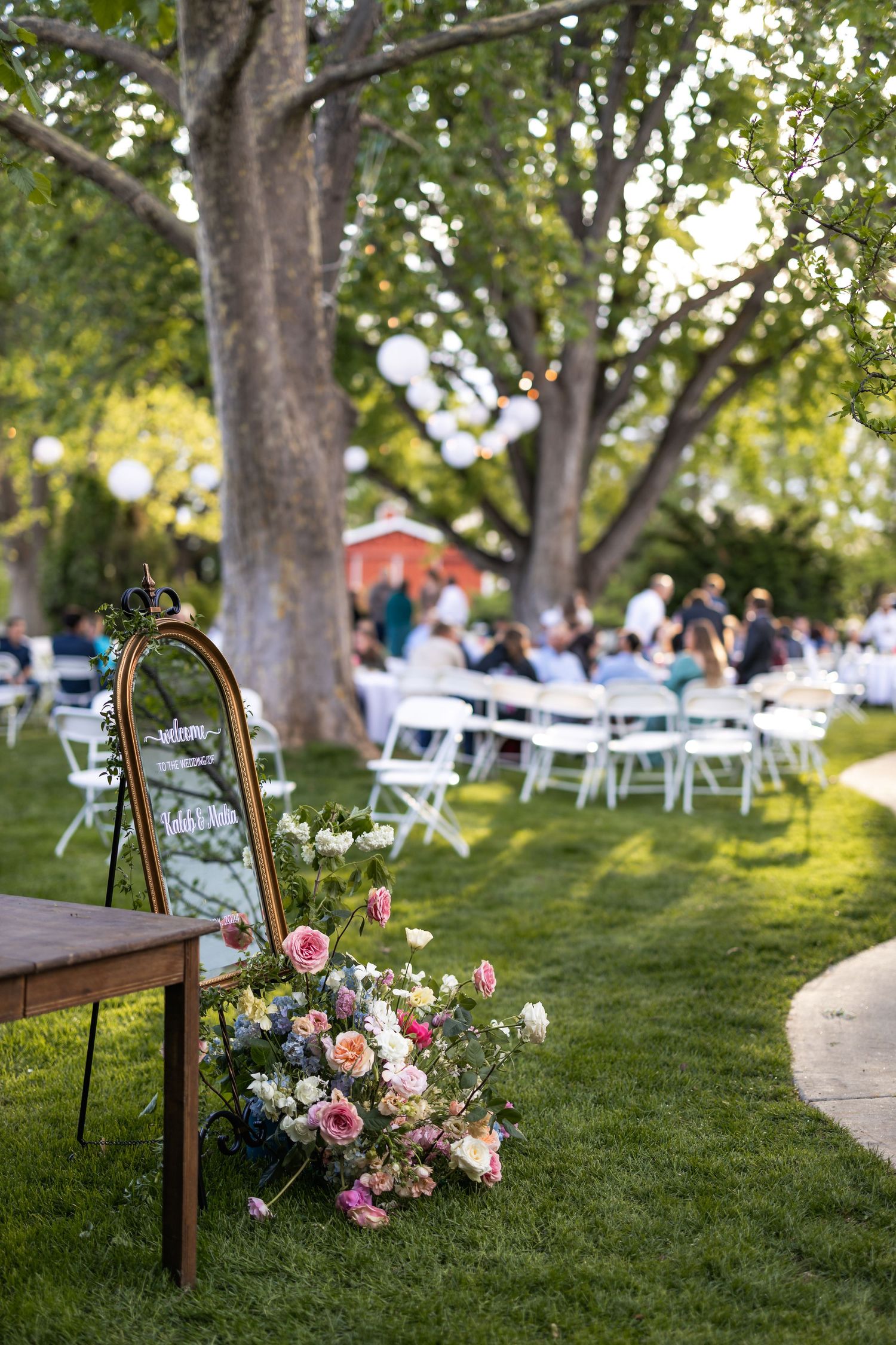 Outdoor wedding reception setup with vintage mirror, pink floral arrangements and white chairs under trees.