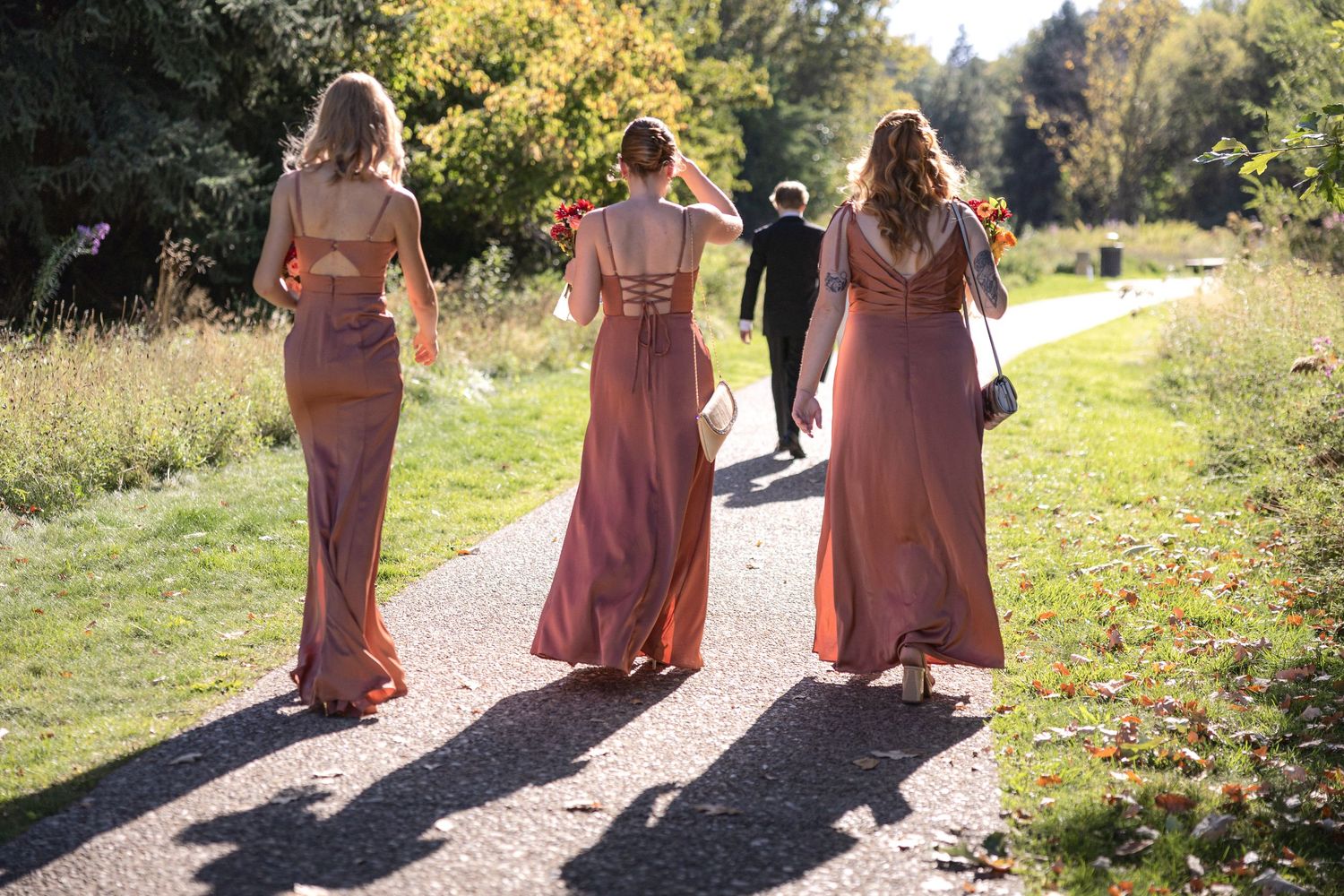 Bridesmaids in coral dresses walking along an outdoor path.