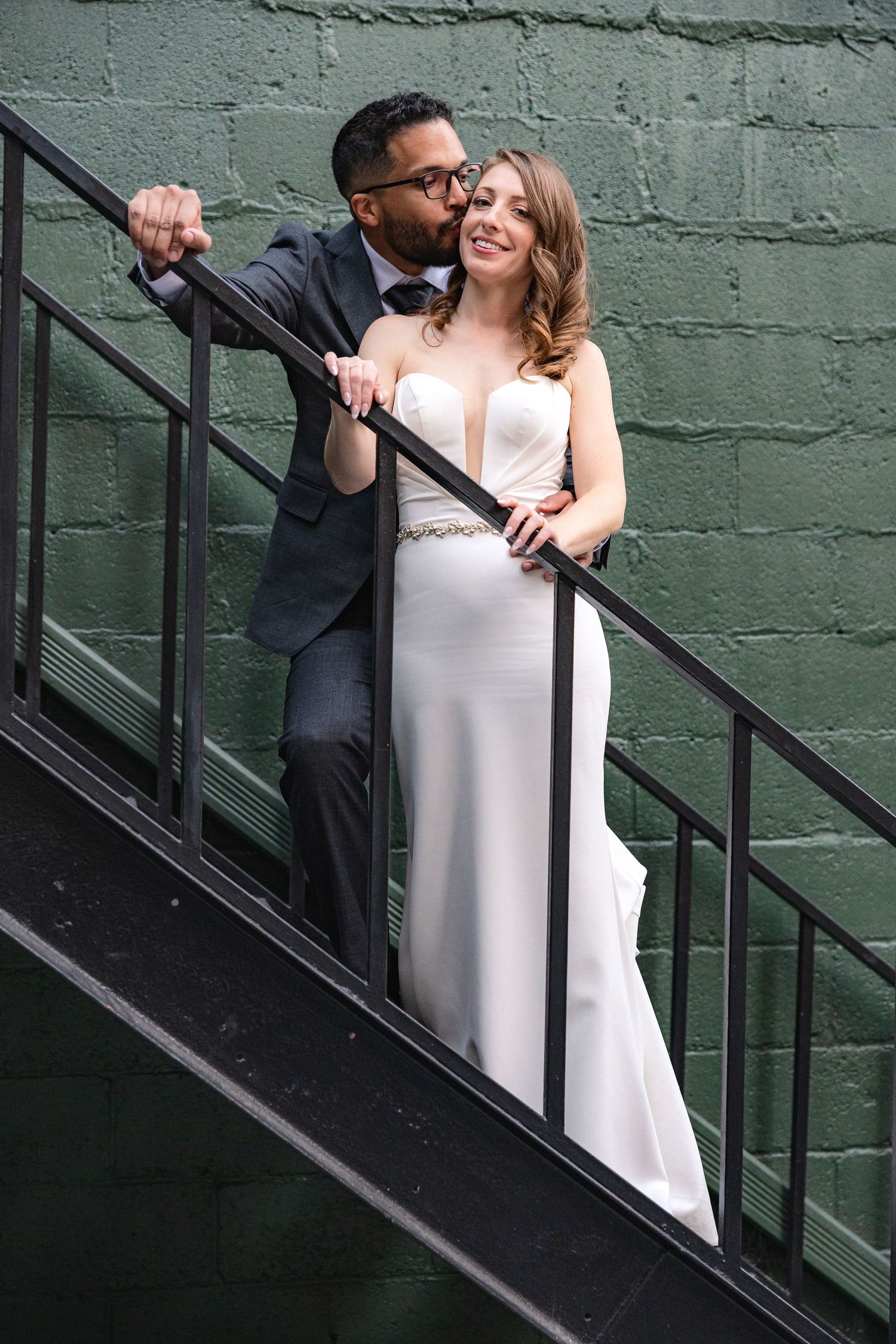 A couple shares a romantic moment on a metal staircase against a green wall.