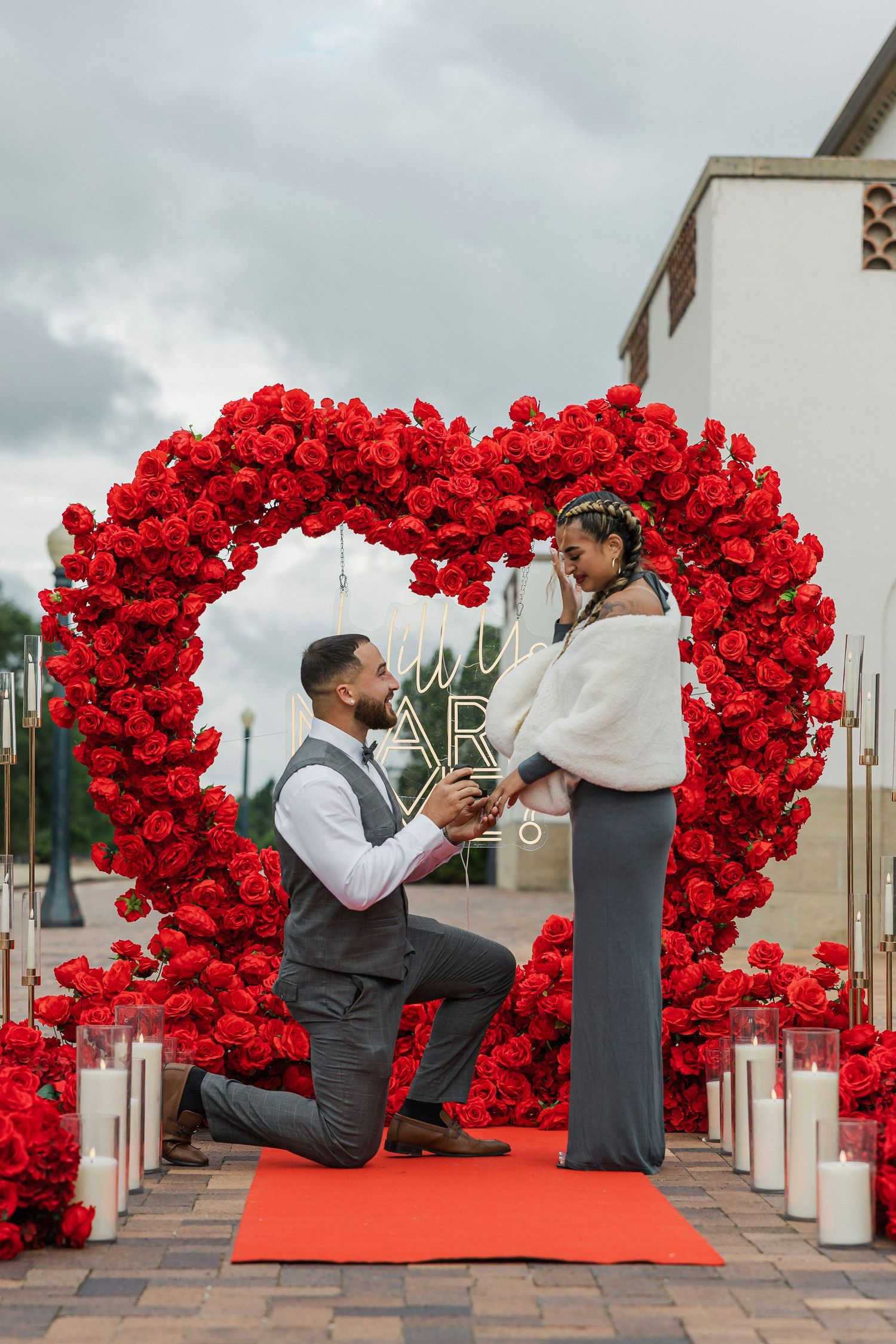 Man proposes on one knee under a large red rose heart arch with candles, as woman reacts with joy outdoors.